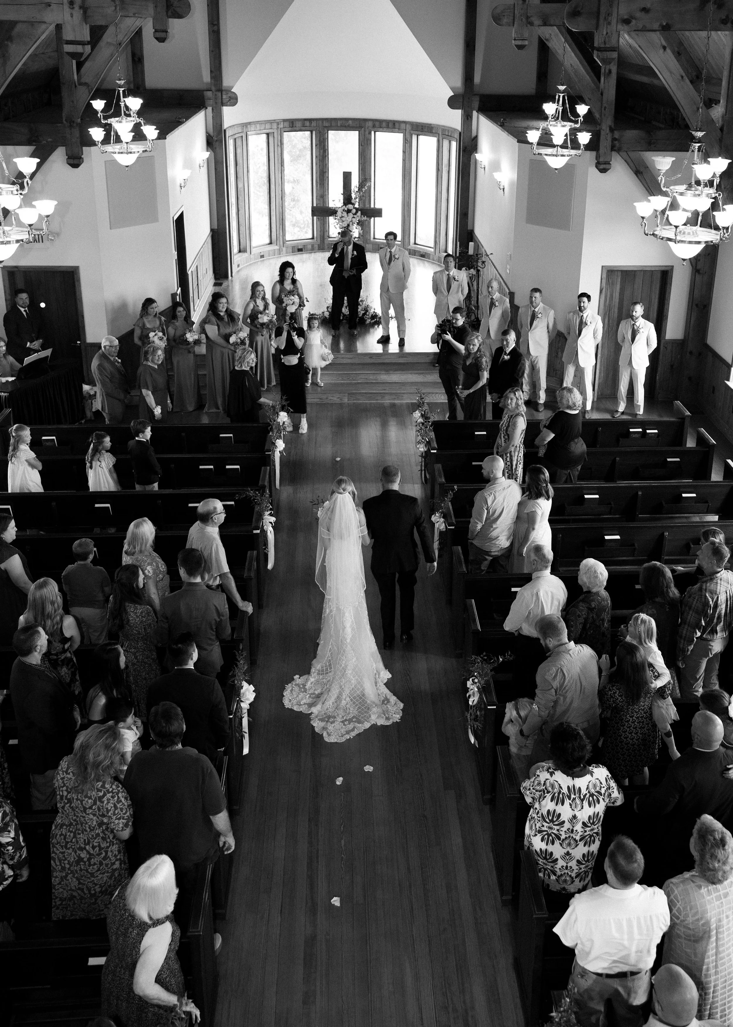 A wedding ceremony inside a church with a bride walking down the aisle toward the altar, which has a large cross and flower arrangements, with guests seated and standing, and wedding party members waiting.