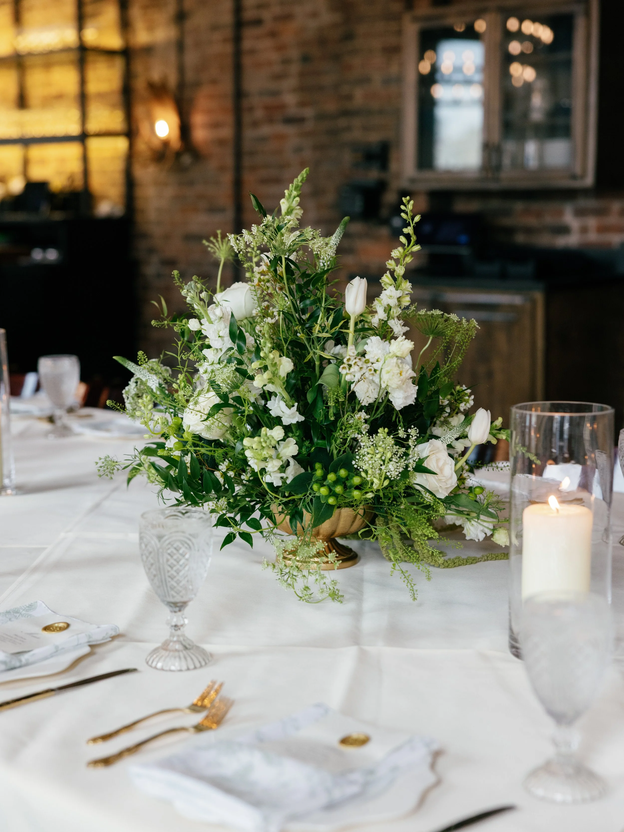 A table decorated with a white tablecloth, a glass vase with a white floral centerpiece, a white candle in a glass holder, and gold utensils.