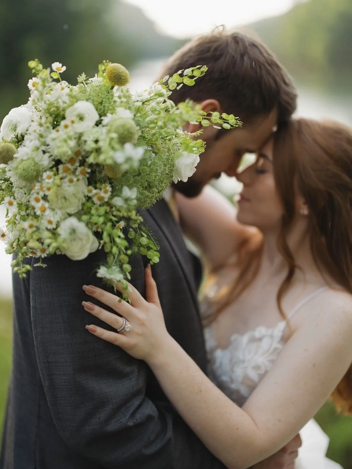 A couple on their wedding day embracing with their foreheads touching, close-up of the bride's hand on the groom's shoulder showing her wedding ring, outdoor setting with a blurred green background, and a large bouquet of white flowers.