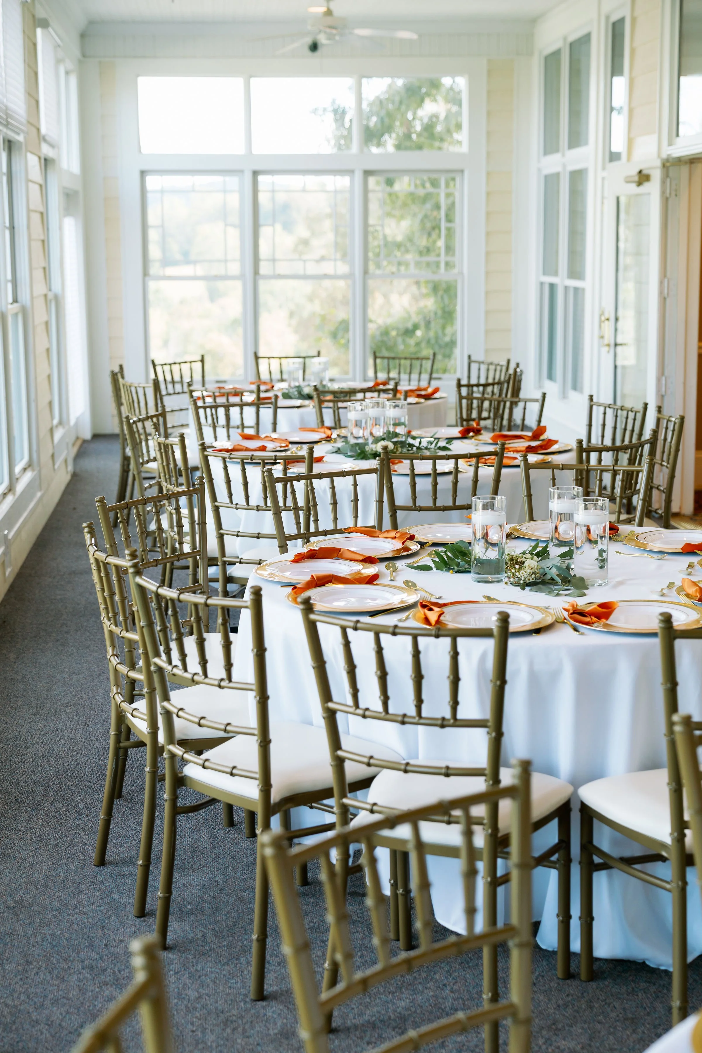 Round tables set for a meal with white tablecloths, gold plates, orange napkins, and glasses of water, arranged in a sunlit room with large windows and gold chairs.