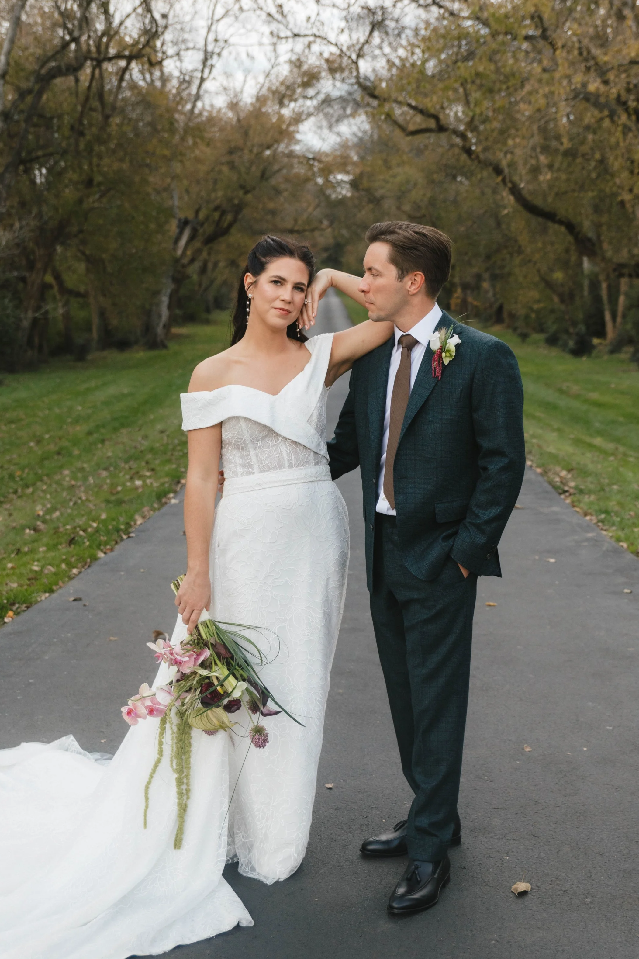 A bride and groom standing on a paved road lined with trees in autumn. The bride is holding a bouquet of pink and purple flowers, wearing a white off-the-shoulder wedding dress. The groom is dressed in a dark suit with a white shirt, brown tie, and boutonniere. The bride has dark hair and is looking at the camera with her hand resting on her head; the groom is looking at the bride with one hand in his pocket.