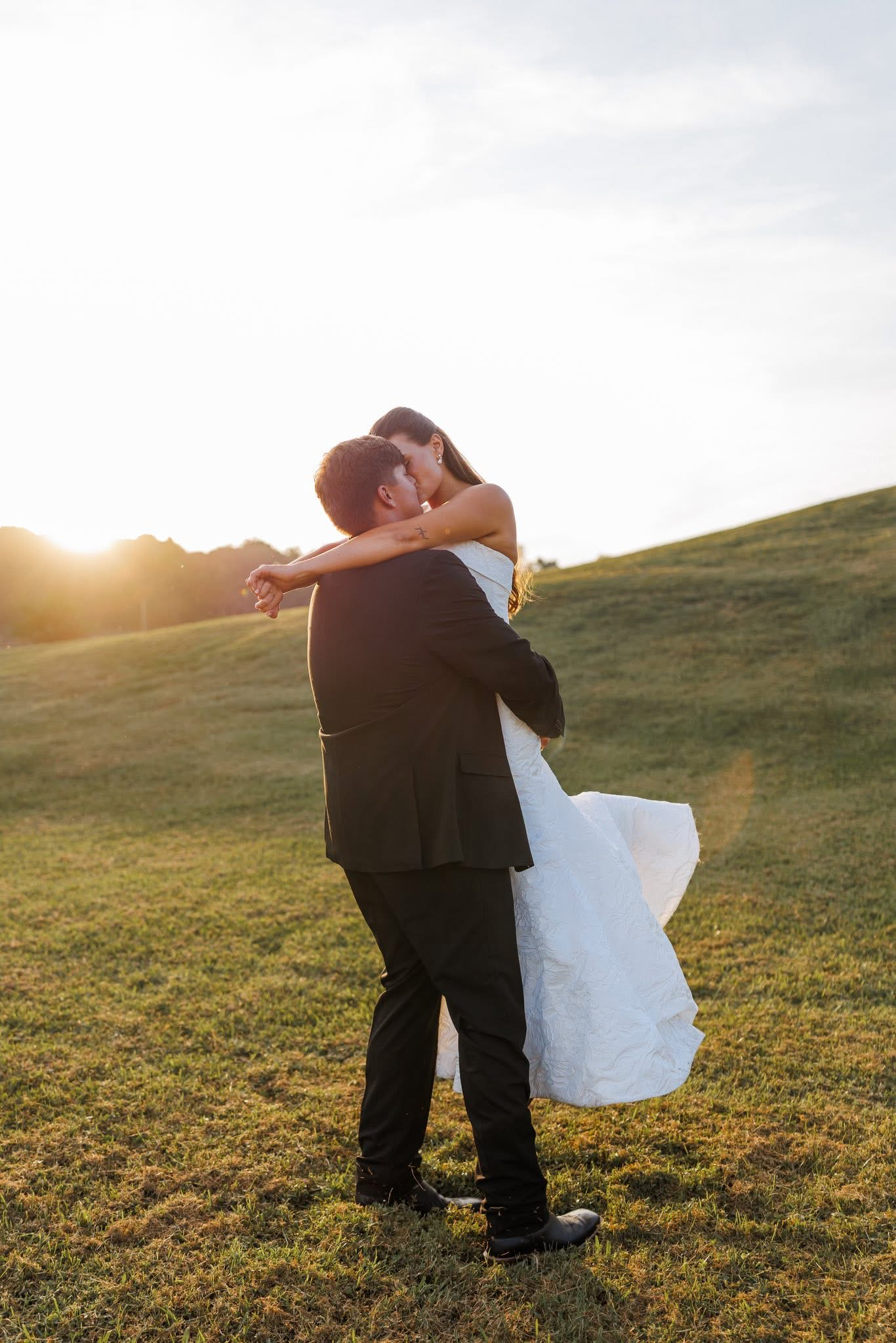 A man in a suit holding a woman in a wedding dress in a field at sunset, sharing a kiss.