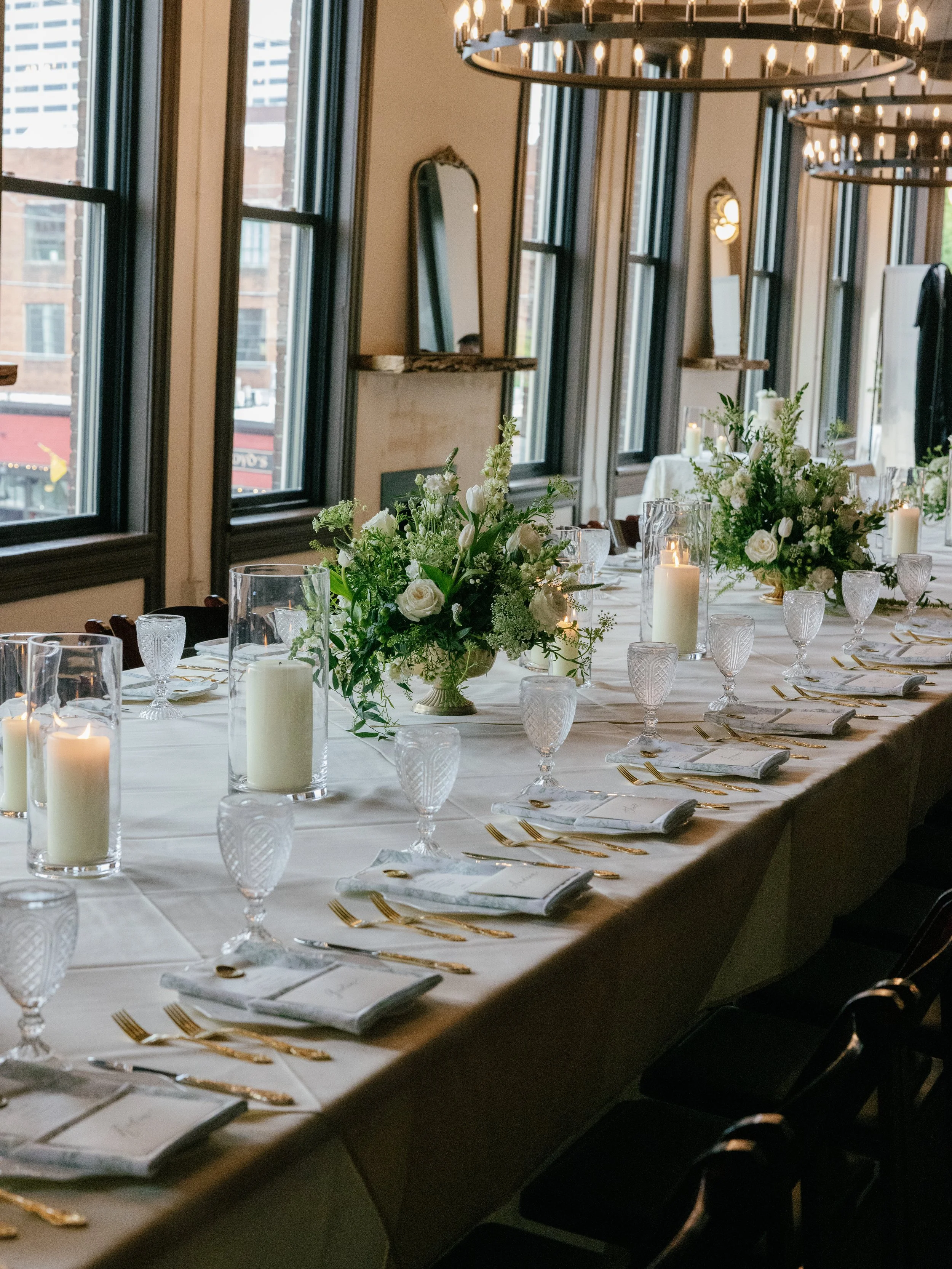 A long banquet table decorated with white flowers, candles, and glassware, set for a formal event inside a room with large windows and hanging circular chandeliers.