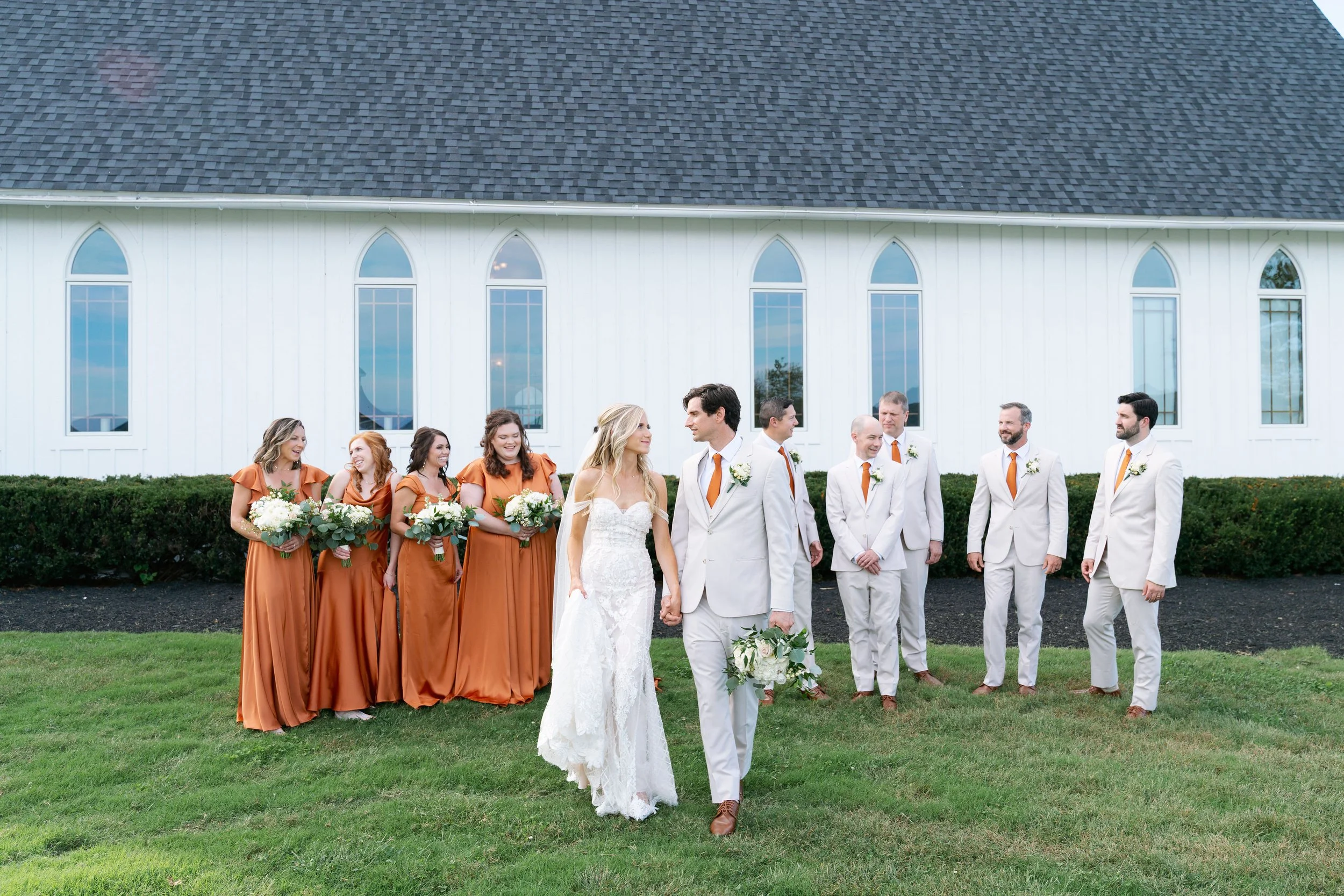 Group of people, wedding party, standing outdoors in front of a white church with tall arched windows, dressed in formal attire with some holding bouquets, celebrating a wedding.