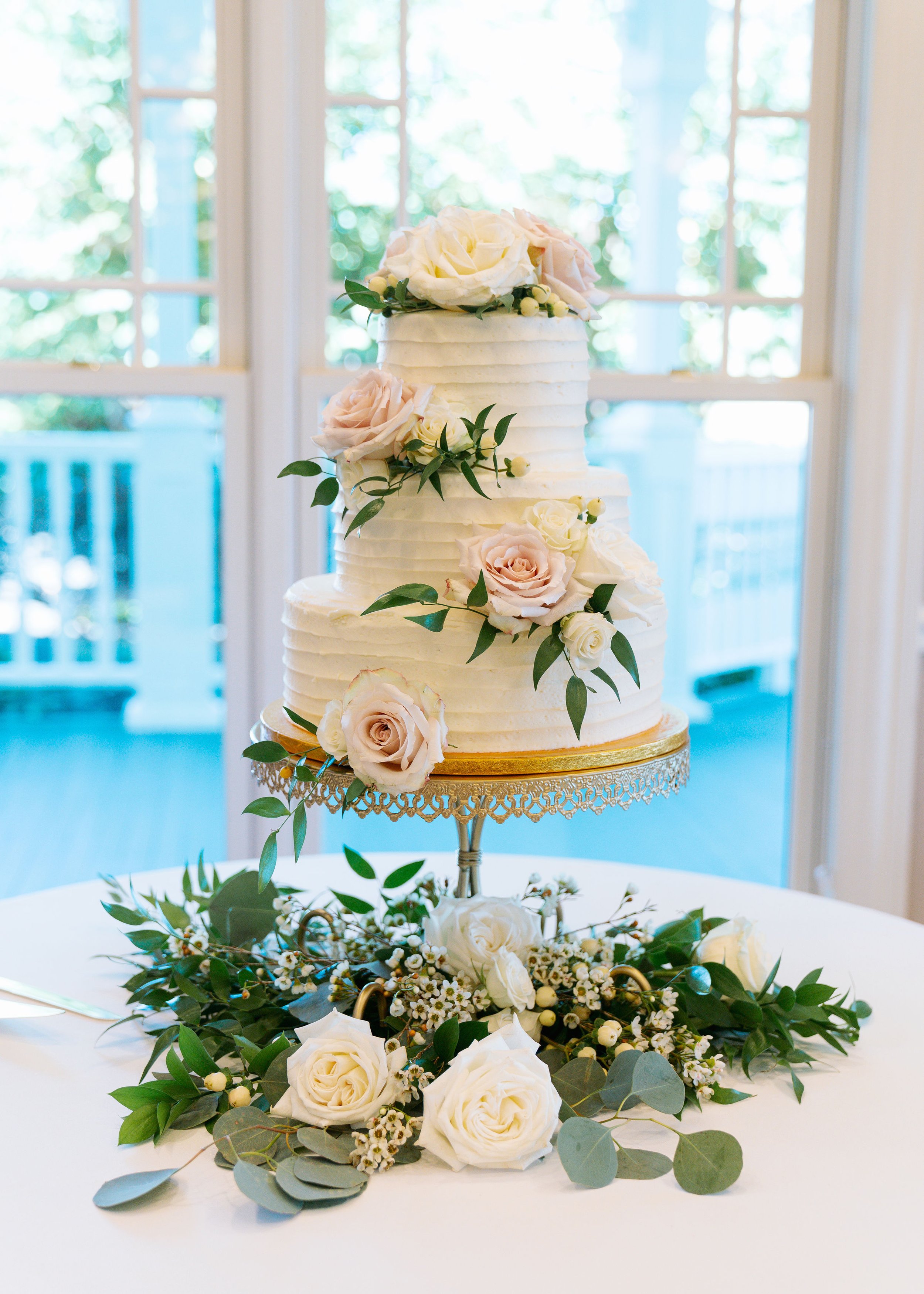 A tiered wedding cake decorated with white and blush pink roses, green leaves, and berries, displayed on a silver stand with floral arrangements and greenery on a white table.