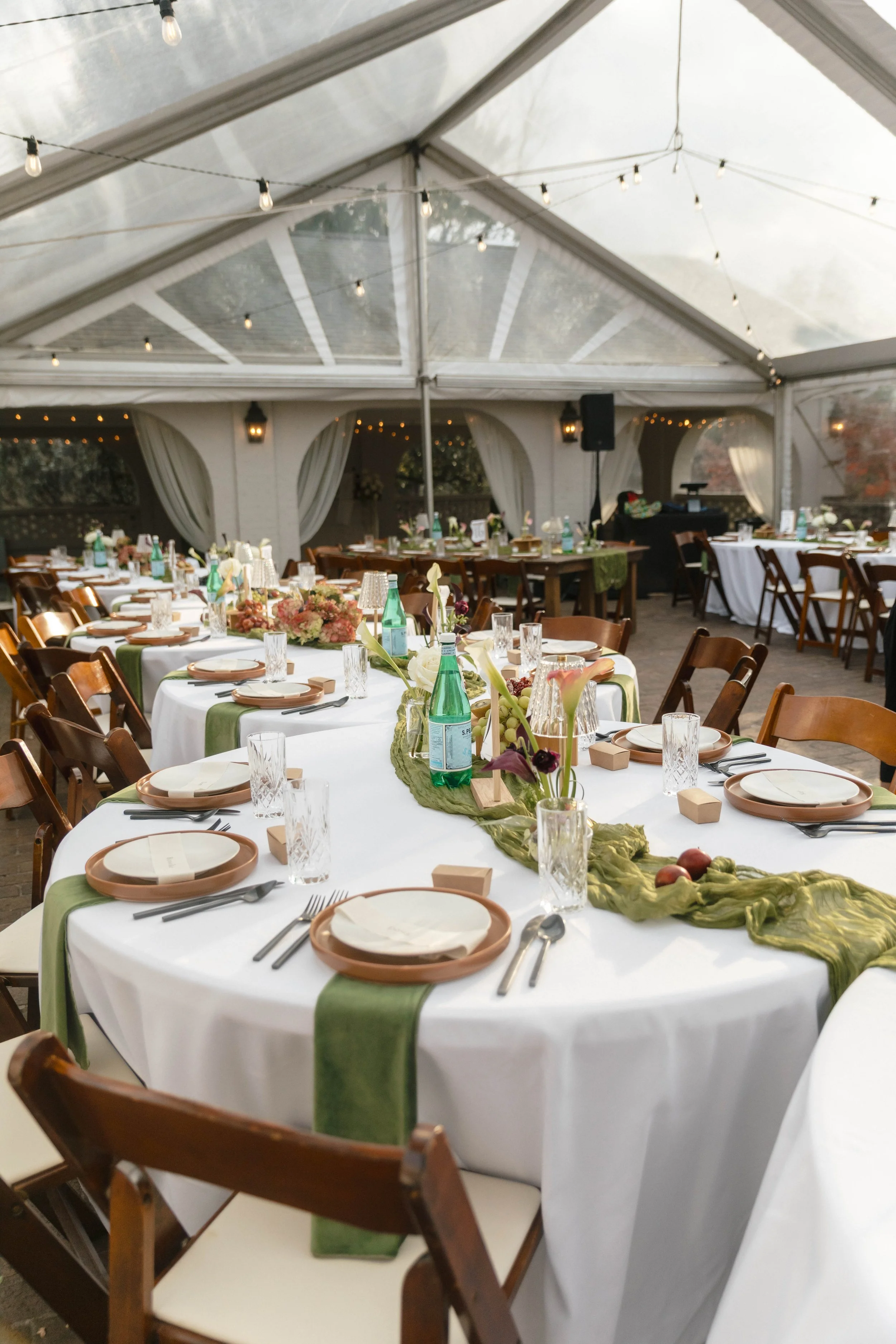 Indoor event space with round tables decorated with green and white table runners, floral arrangements, water bottles, glassware, plates, and silverware. Tent with string lights overhead and curtains along the sides.