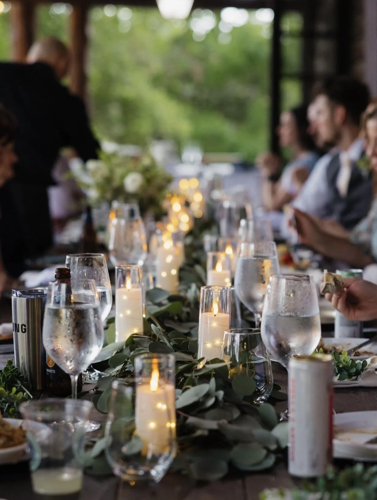 A candlelit dinner table at a formal event with glasses of water, greenery, and blurred guests in the background.