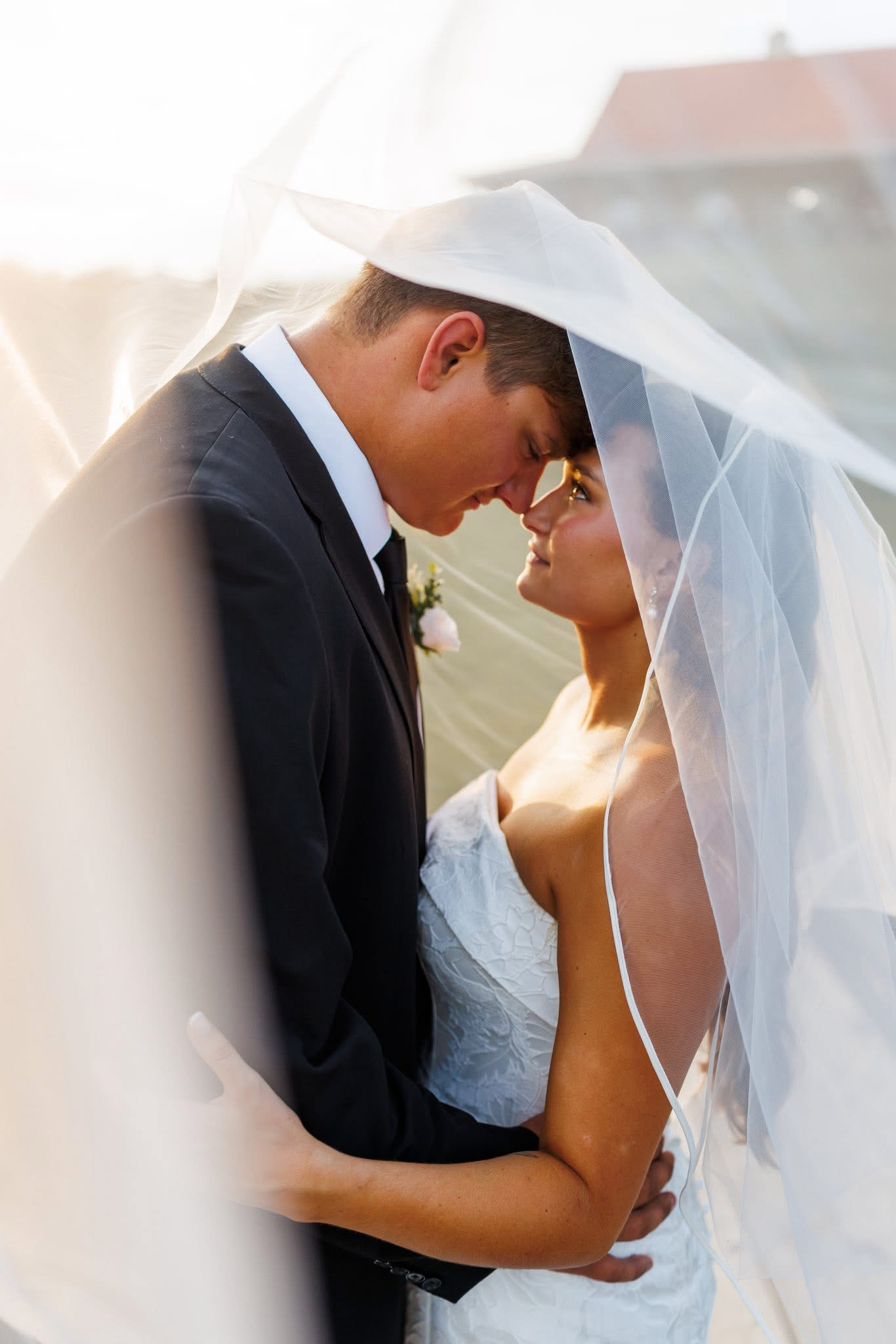 A bride and groom with foreheads touching, smiling, outdoors in sunlight, bride wearing veil and white dress, groom in tuxedo, intimate wedding moment.