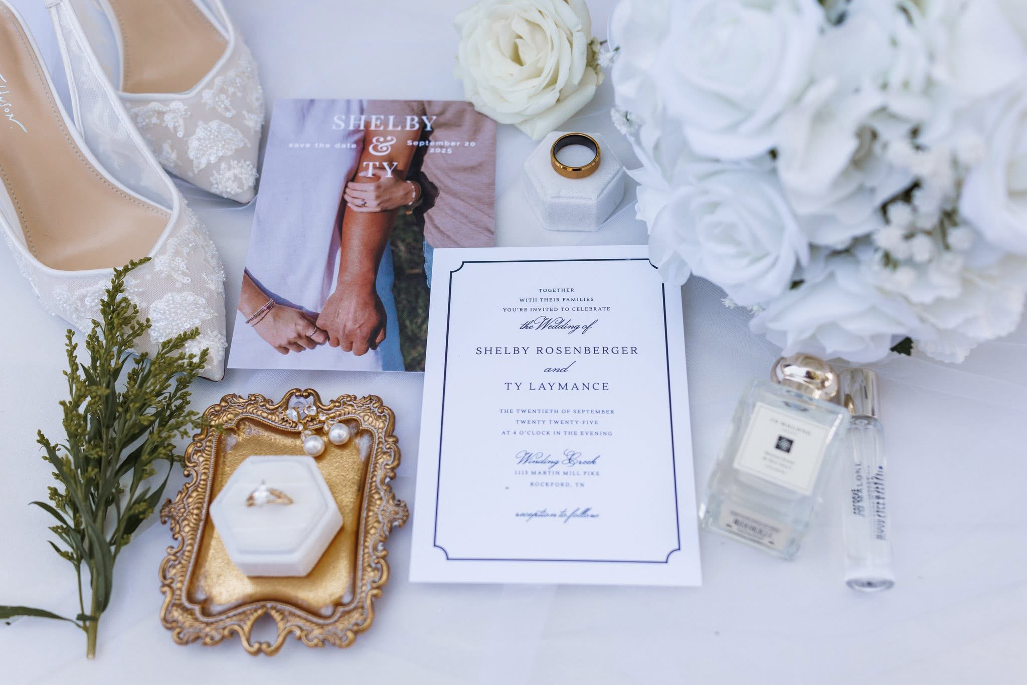 A wedding invitation surrounded by white roses, a gold wedding band, a perfume bottle, and jewelry, with a pair of bridal shoes and a white flower bouquet in the background.