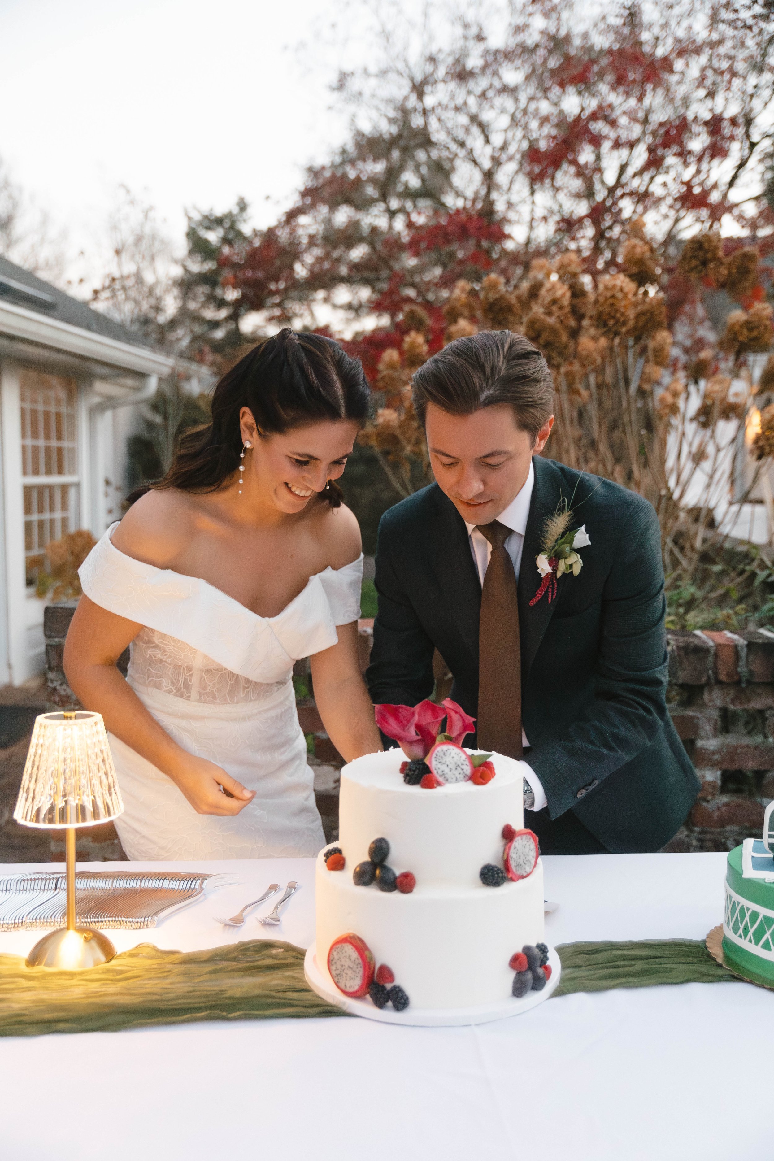 A bride and groom cutting a wedding cake outdoors during sunset. The cake is decorated with berries and dragon fruit. The scene includes a table with utensils and a table lamp, with autumn trees in the background.