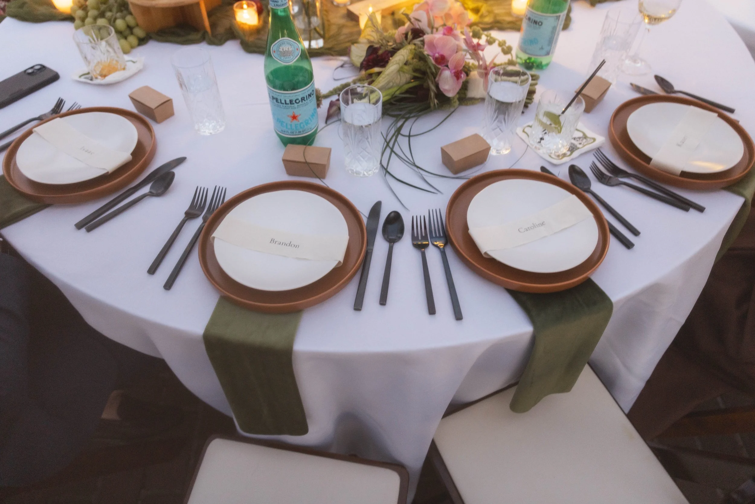 A round banquet table decorated with a white tablecloth and olive green napkins. Place settings include brown plates, white napkins, and black cutlery. The table features water bottles, glasses, floral centerpiece, and small gift boxes.
