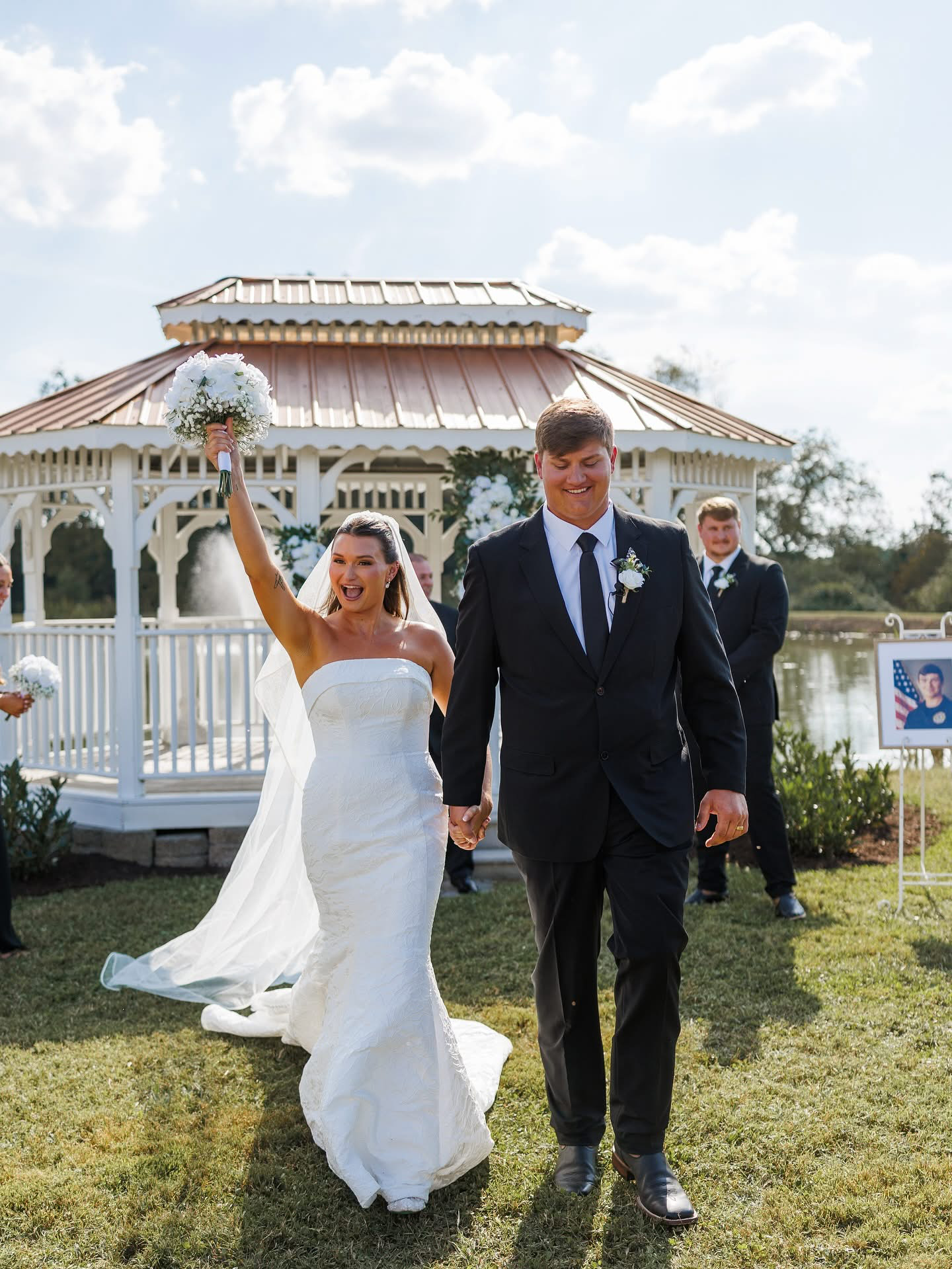 A newlywed couple holding hands walks outdoors during wedding celebration. The bride holds a bouquet in her right hand, raised in the air, smiling and wearing a strapless white wedding dress. The groom, in a black suit with a white shirt and black ti