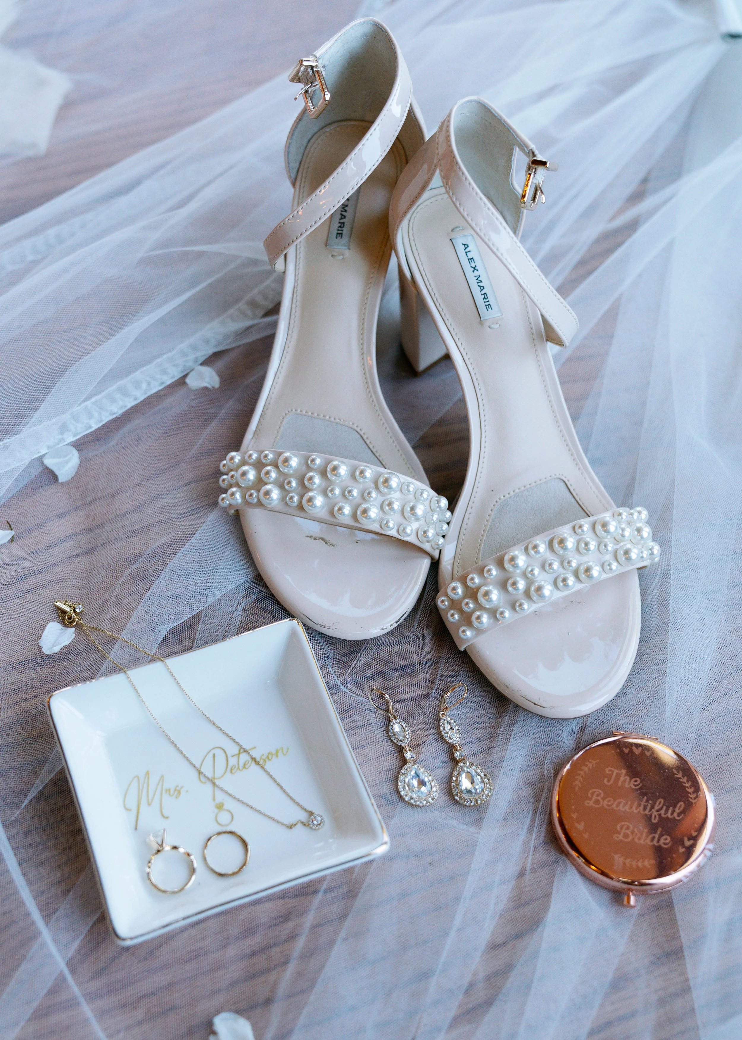 A pair of white high heel shoes with pearl embellishments on the toes, placed on a soft, sheer fabric. Surrounding the shoes are jewelry pieces, a small white box with gold lettering, and a round container with the words 'The Beautiful Bride' on it.