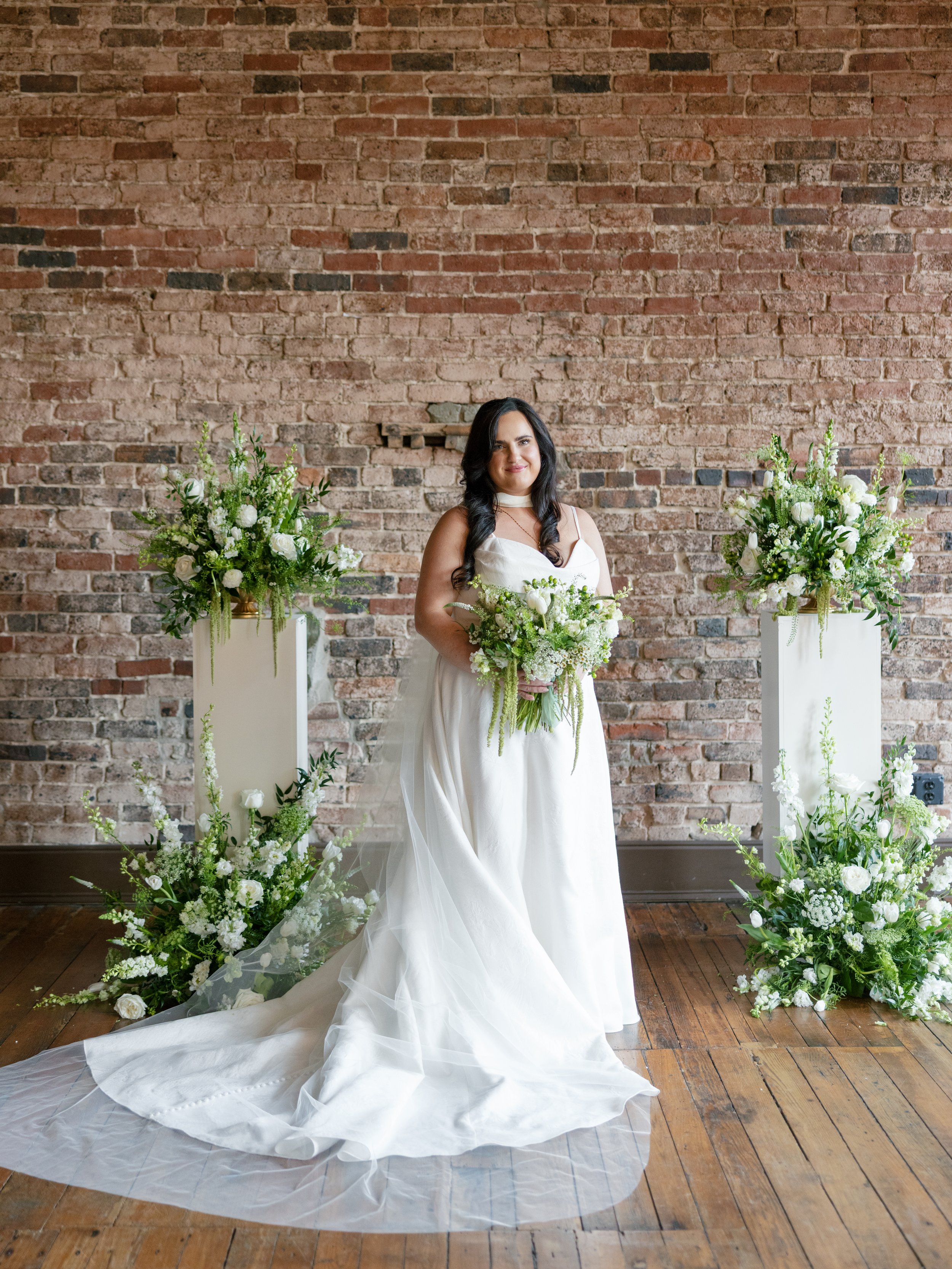 A woman in a white wedding dress holding a bouquet, standing in front of a brick wall with surrounding floral arrangements.