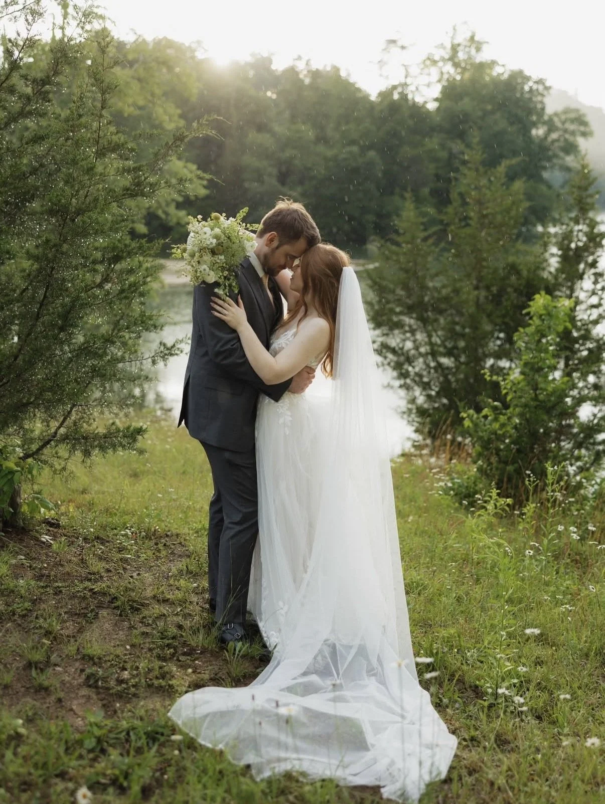 A bride and groom embrace outdoors near a river, with greenery and trees in the background, during their wedding photoshoot in the late afternoon.