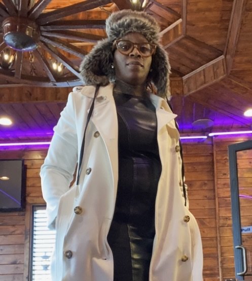 A woman wearing a beige trench coat, black leather dress, glasses, and a warm fur hat standing indoors with a wooden ceiling and purple lighting in the background.