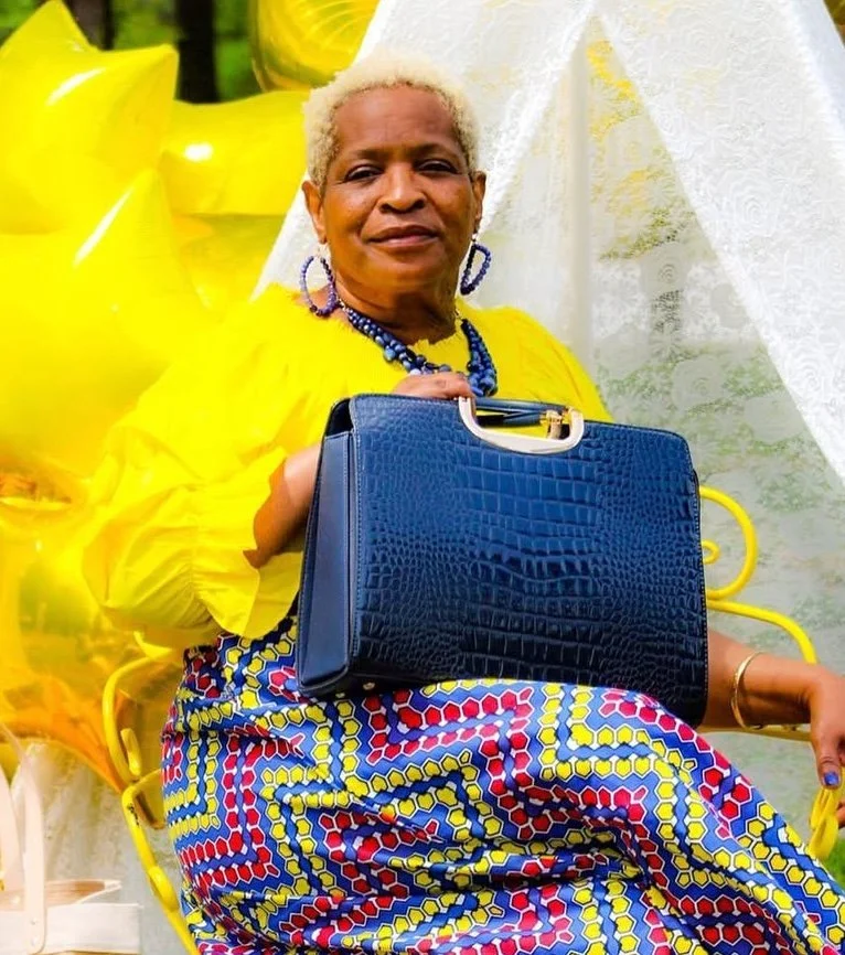 An elderly woman with short platinum blonde hair, wearing a bright yellow top, vibrant patterned skirt, blue accessories, and holding a blue handbag. She is sitting outdoors in front of a white structure with yellow balloons around her.
