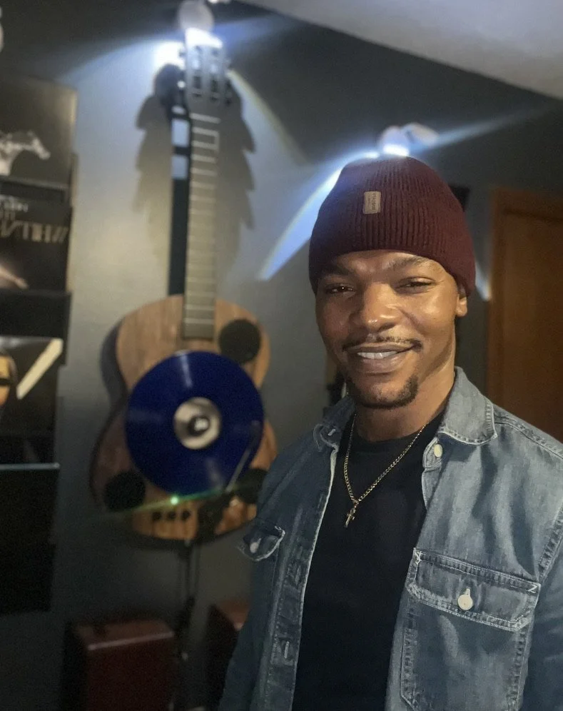 A man wearing a red beanie, a black shirt, and a denim jacket taking a selfie in front of a guitar wall decoration with a CD attached to the guitar.