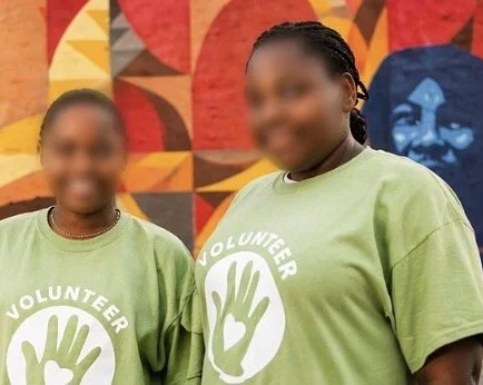 Two women wearing green volunteer t-shirts with hand graphics, standing in front of a colorful mural.
