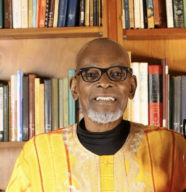 A smiling Black man with glasses, gray beard, wearing traditional yellow African attire, standing in front of a bookshelf filled with books.