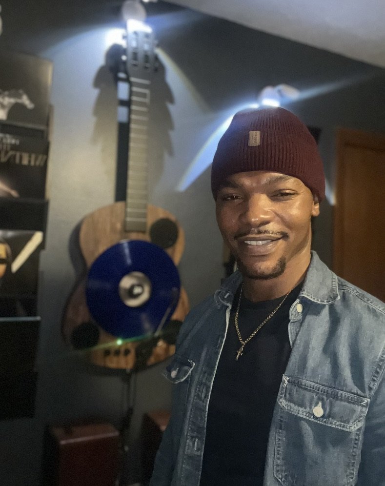 A man wearing a maroon beanie and denim jacket, smiling at the camera, standing in a room with a guitar wall decoration and a black shelf in the background.