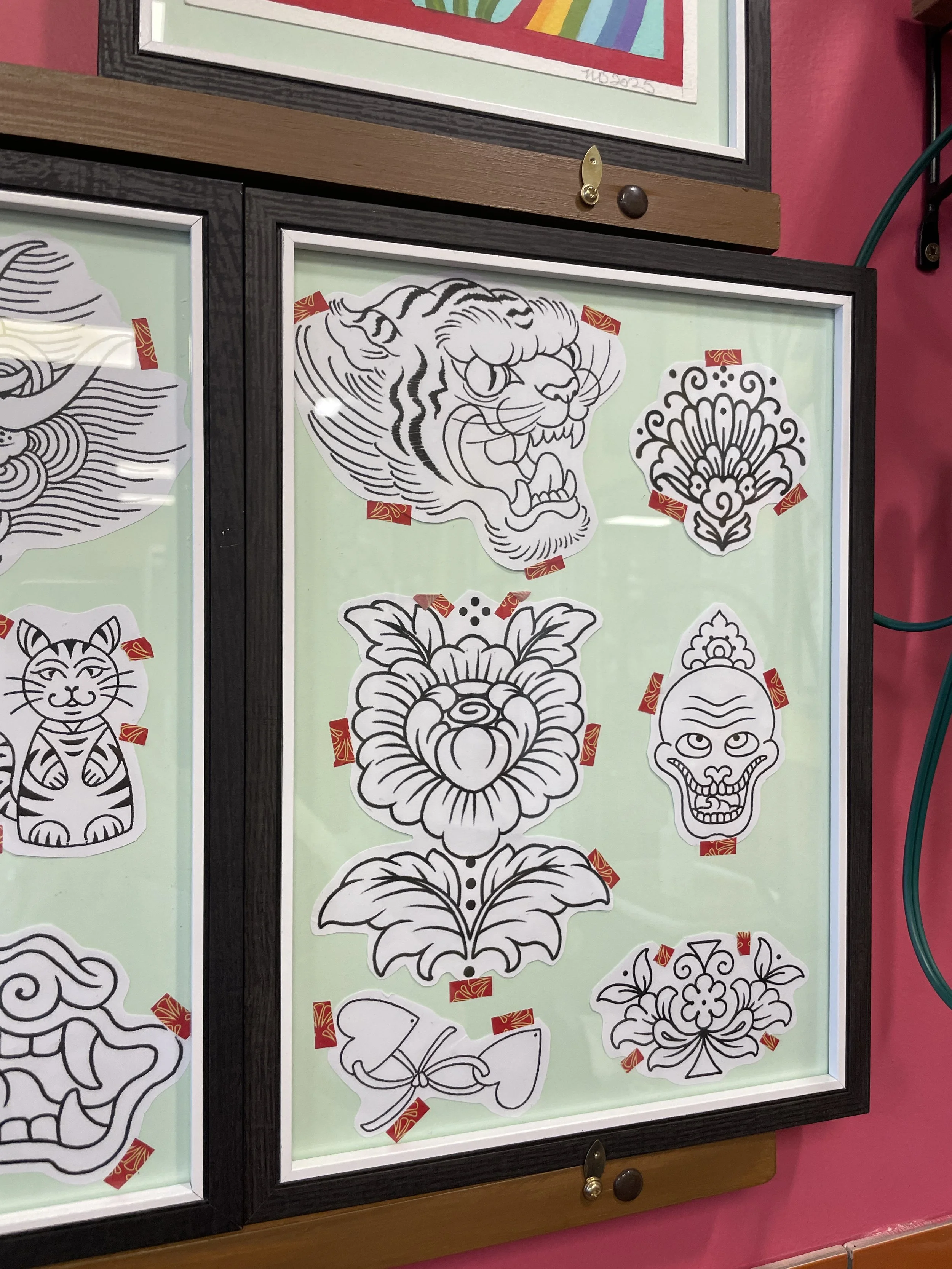 Black and white tattoo-style drawings of various animals and floral designs displayed on a framed glass surface in an indoor setting.