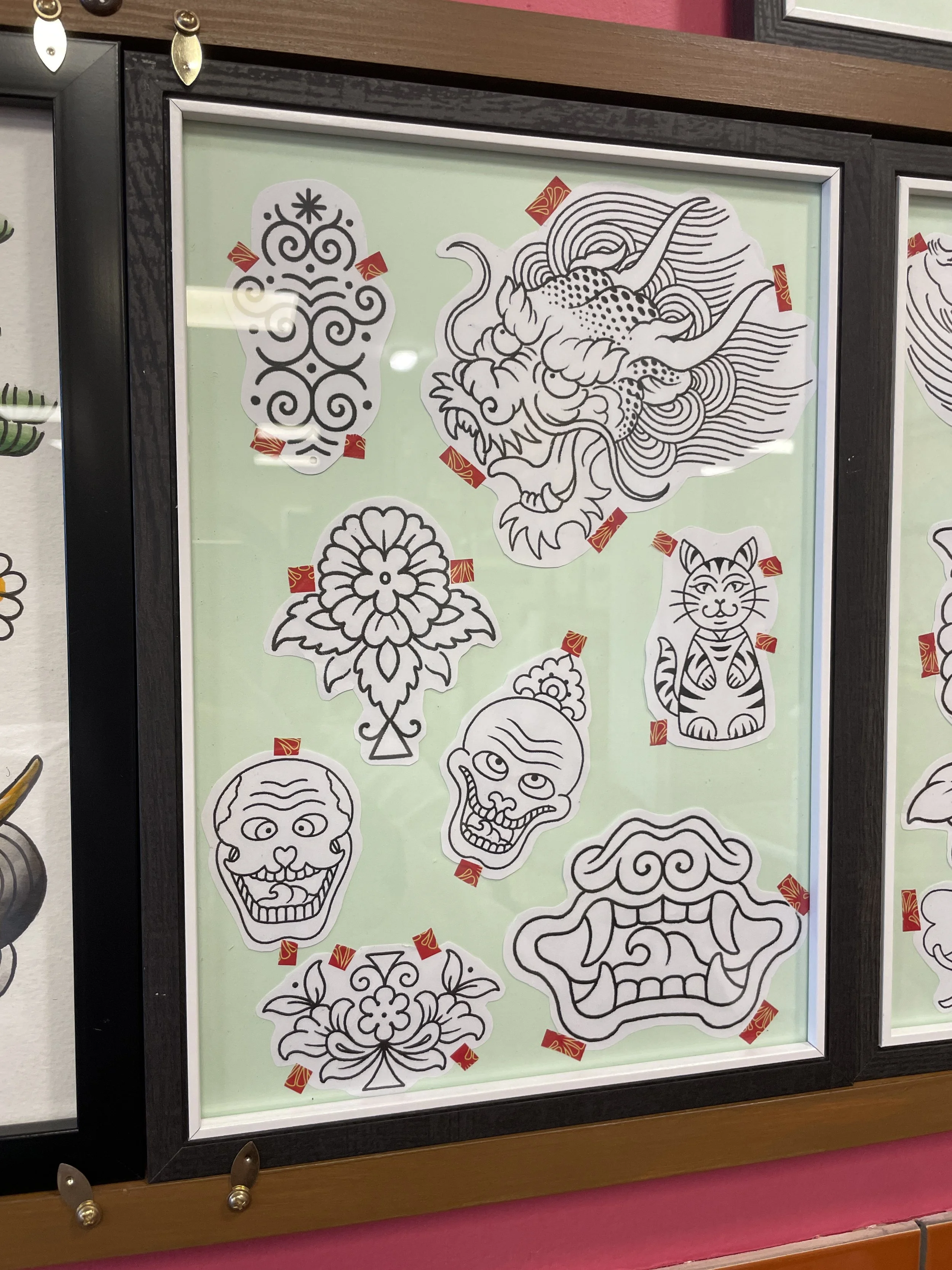 Black and white tattoo flash art designs taped to a light green background, including a detailed lion's head, a stylized face, a skull, a tiger, and other traditional tattoo images.