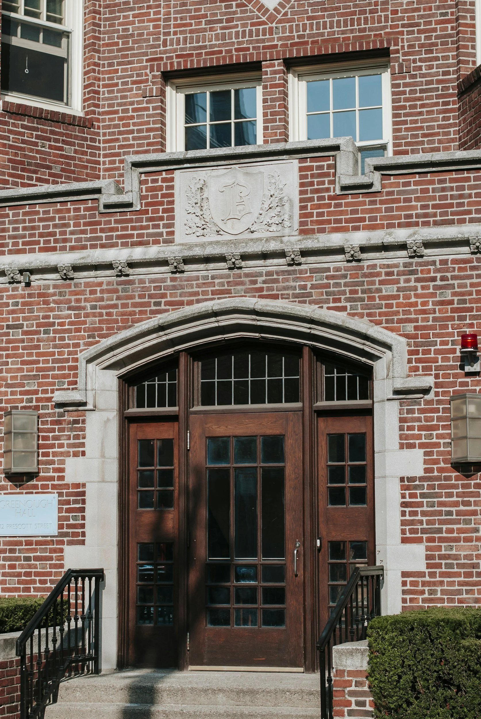 Close-up of a brick building entrance with a wooden door and arched window above, flanked by steps, shrubbery, and black railings.
