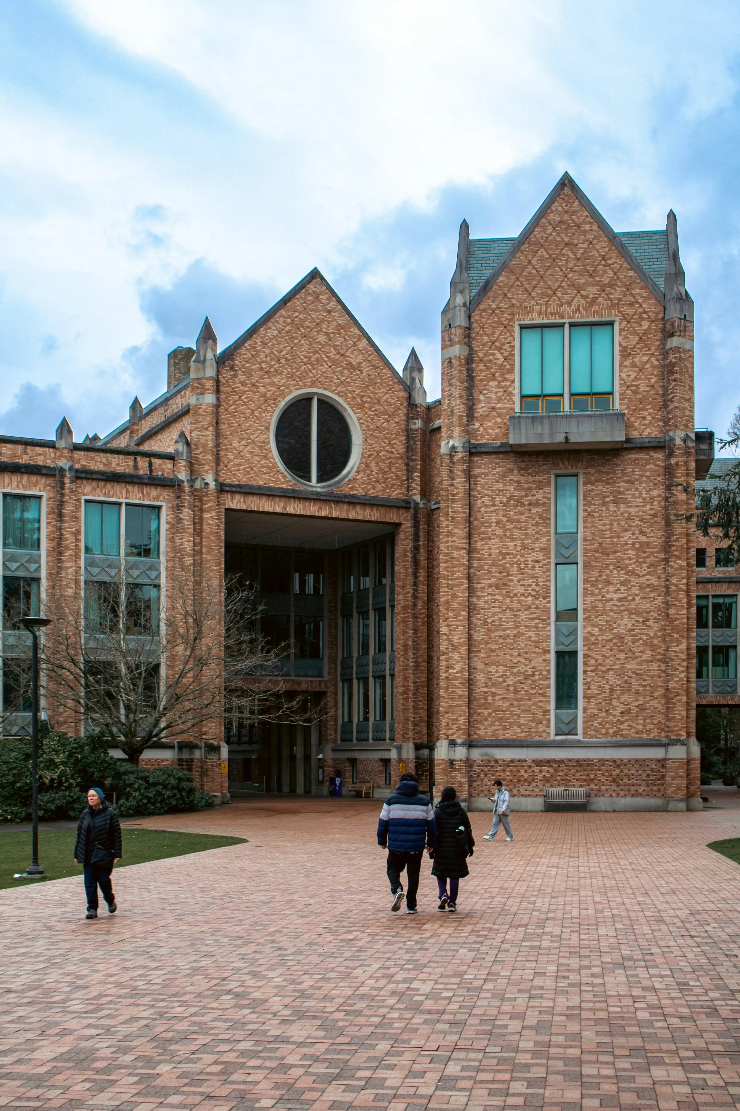People walking on a brick-paved courtyard in front of a large brick building with distinctive peaked roofs and tall windows.