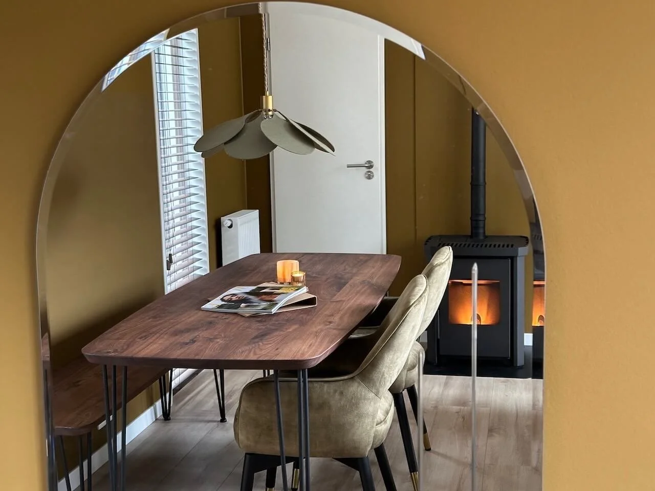 Een eetkamer met een houten tafel, beige stoelen, een grote muur spiegel, een houtkachel met een open deurtje, en een raam met witte jaloezieën.