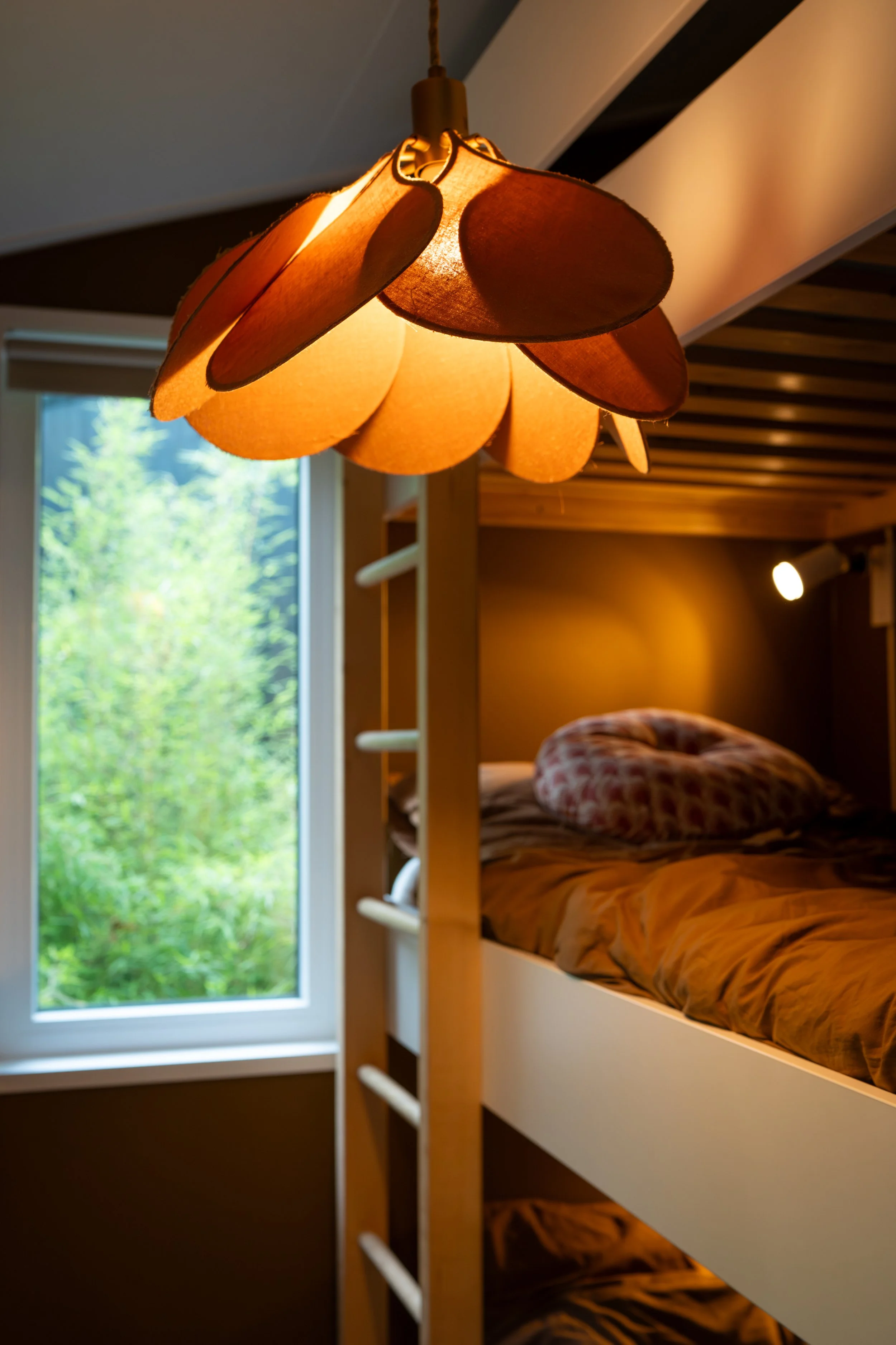 Een houten bed met een gestreept dekbed en een ballonvormig hoofdkussen, naast een grote raam met een groen landschap buiten, en een plafondlamp met een lampenkap van papier of stof.