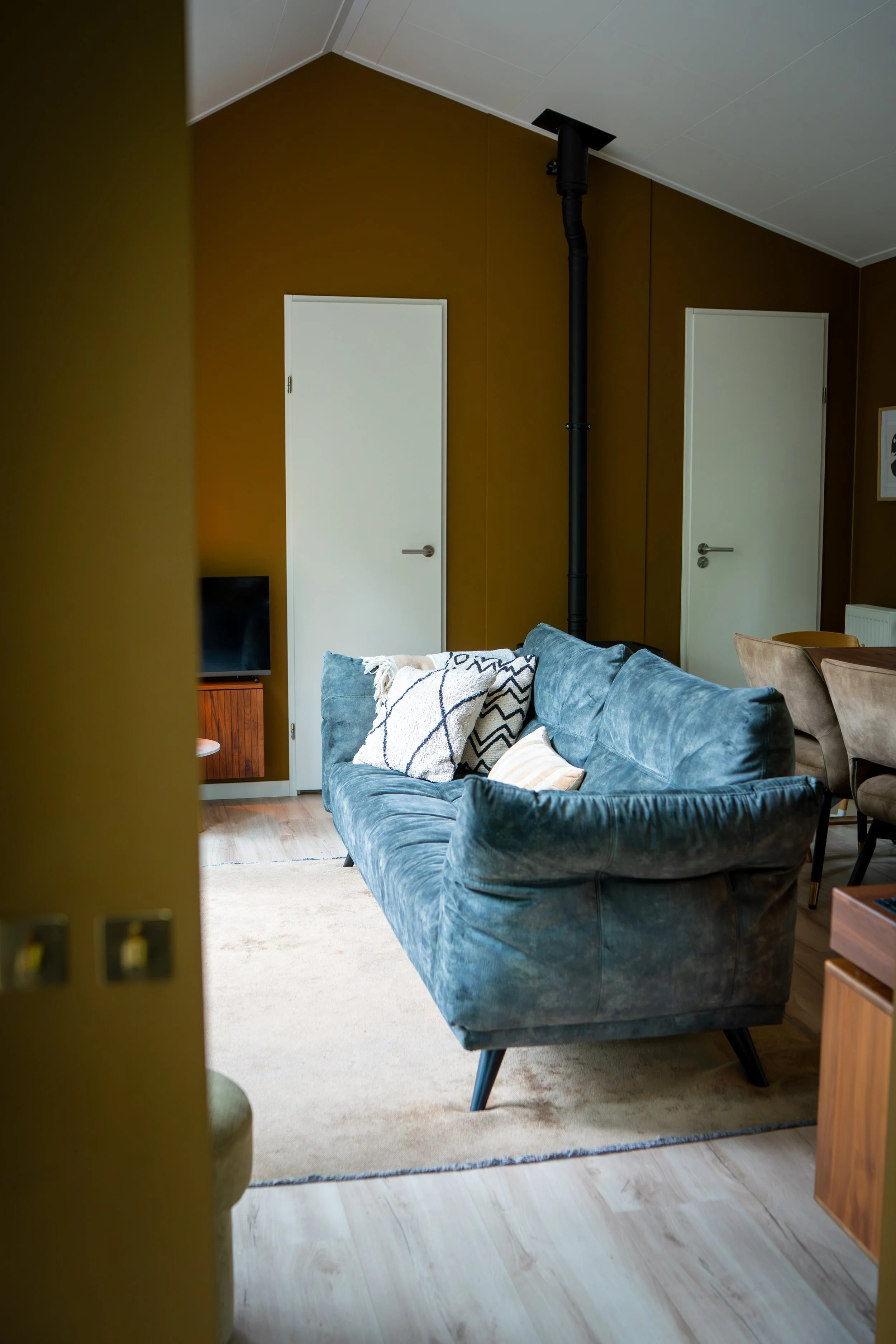 Een woonkamer met een blauwe bank en kussens, een houten vloer en een beige tapijt. Twee witte deuren en een zwarte rookkanaal in de muur.