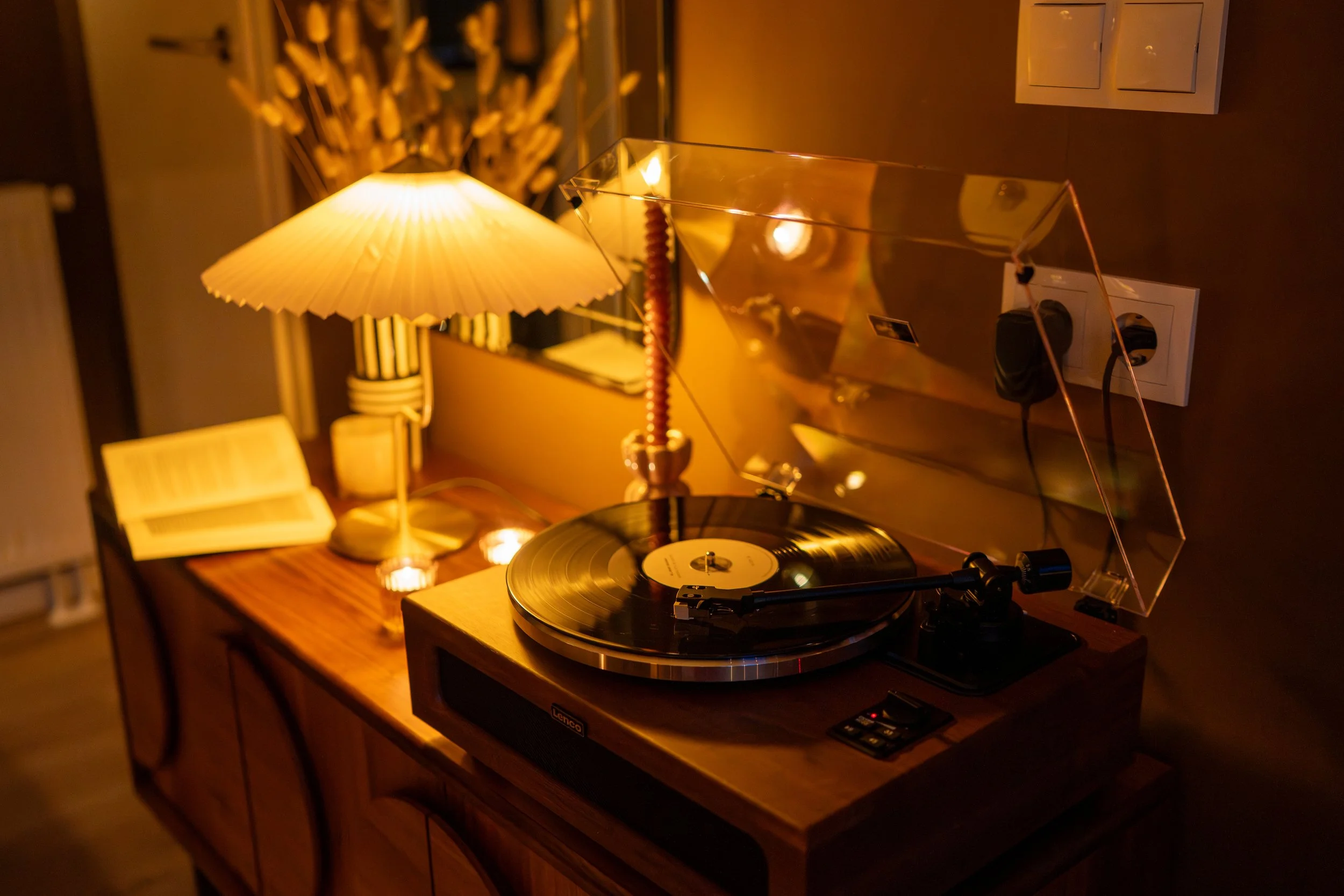 Een draaitafel met een vinylplaat, een lamp met een stoffen kap, een open boek en wat kaarsen op een houten tafel, in een warme, sfeervolle kamer.