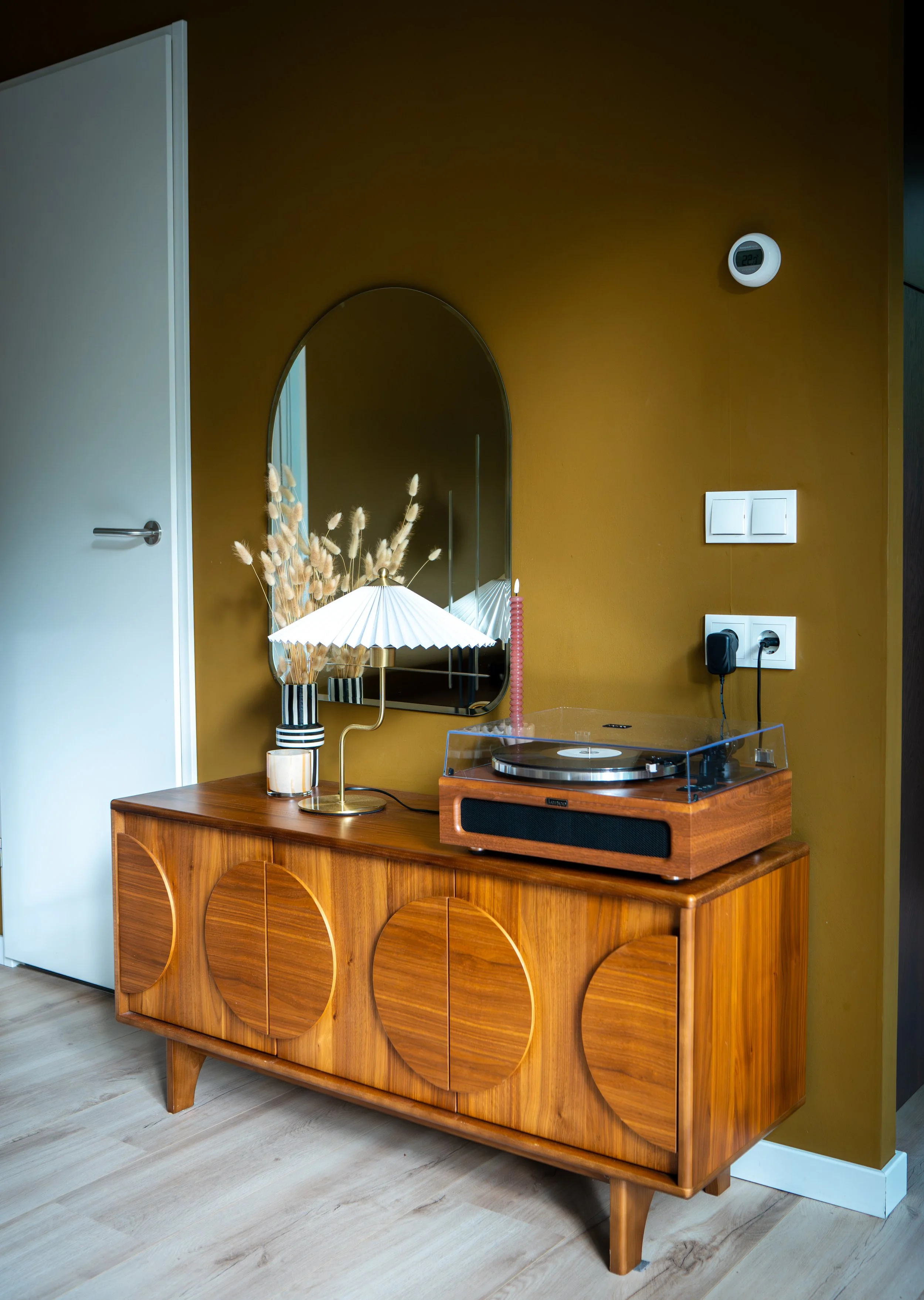 Een houten dressoir met ronde patronen, een tafelklok, een schilderij en een spiegel, geplaatst in een kamer met een blauwe muur en een open witte deur.