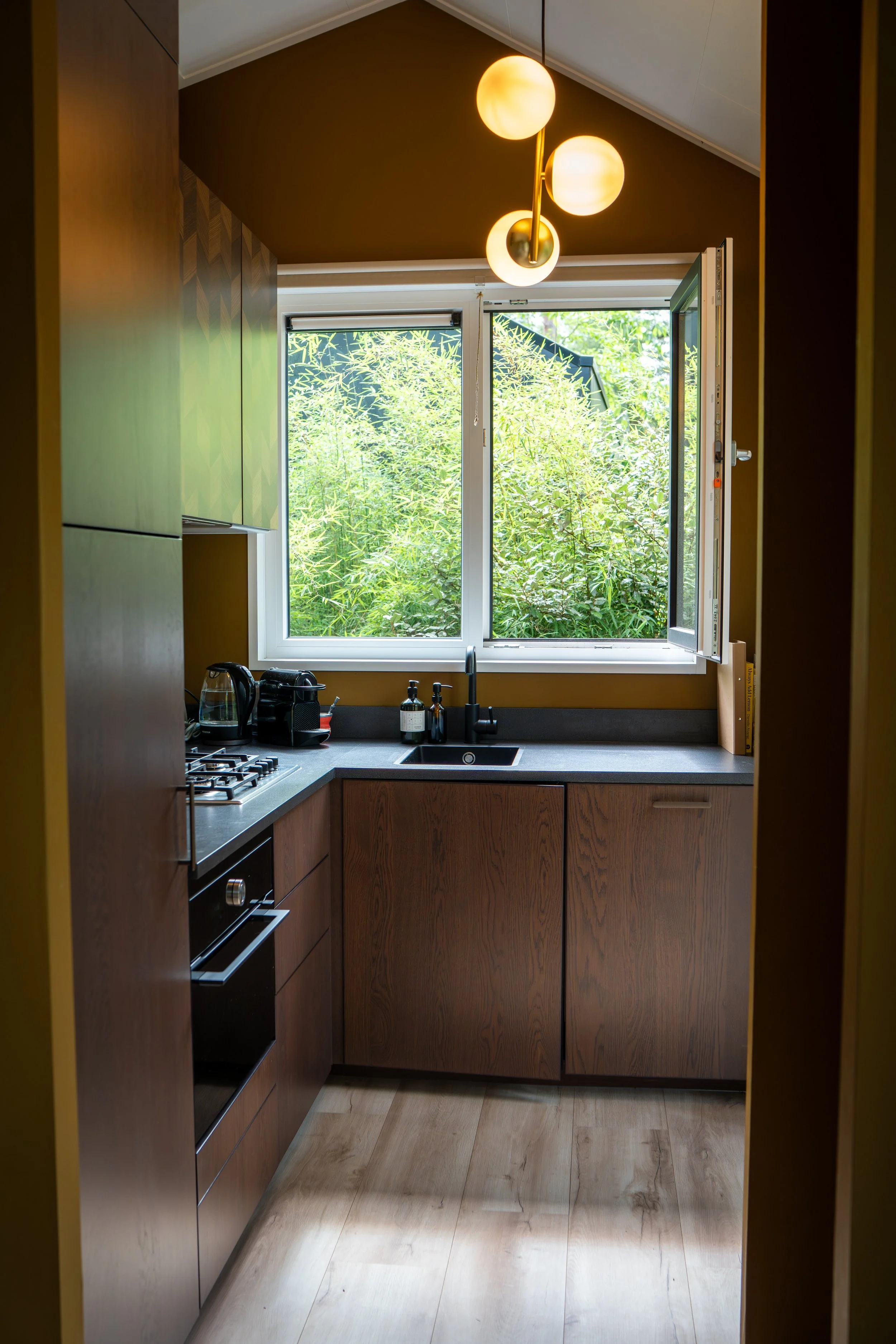 Kleine keuken met houten kasten, een raam met uitzicht op groen, een ingebouwde oven en een wastafel. Boven de keuken hangt een plafondlamp met drie bolvormige lampen.