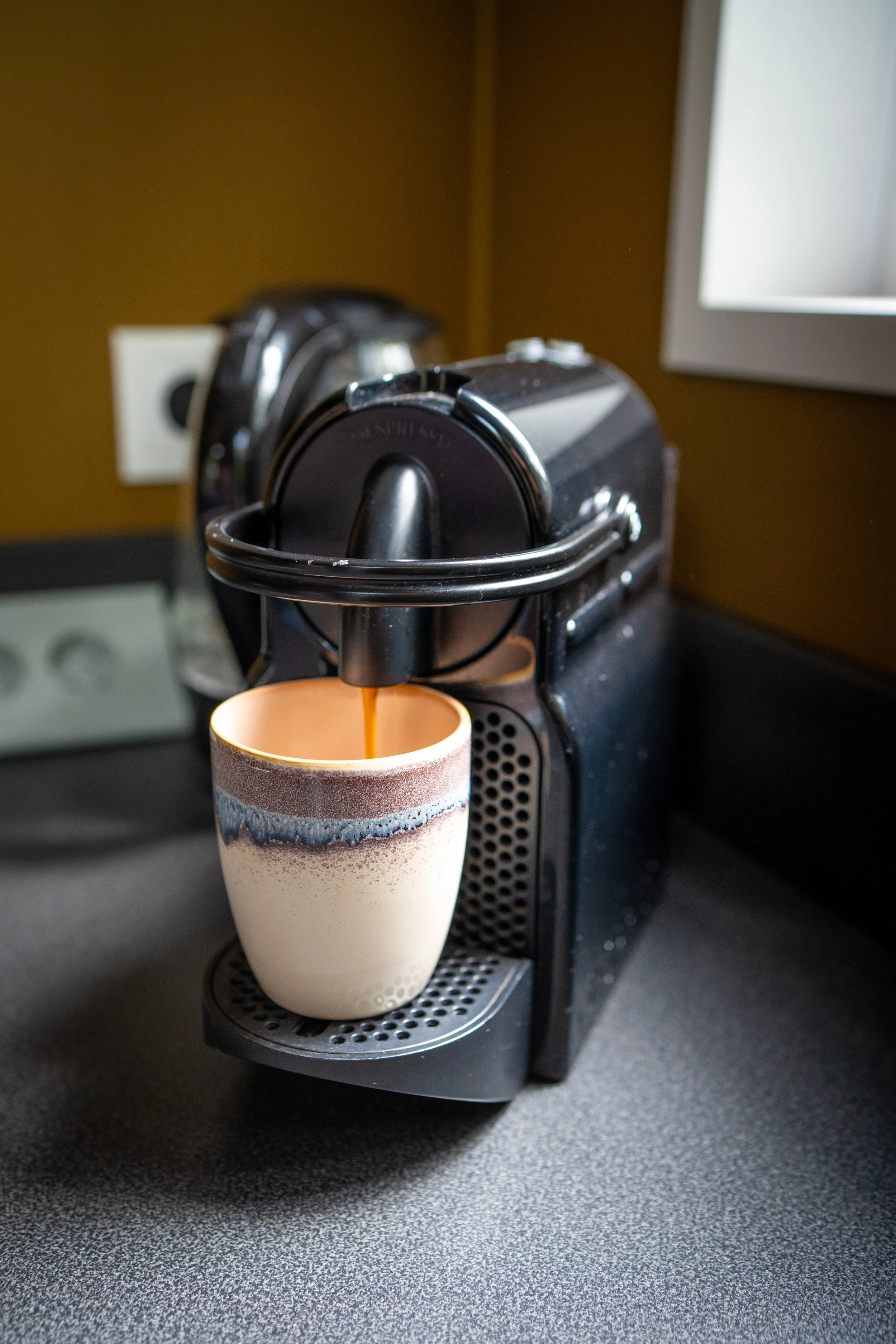Een zwart koffiezetapparaat dat koffie schenkt in een koffiemok met een aardewerk uiterlijk, op een zwarte werkblad.