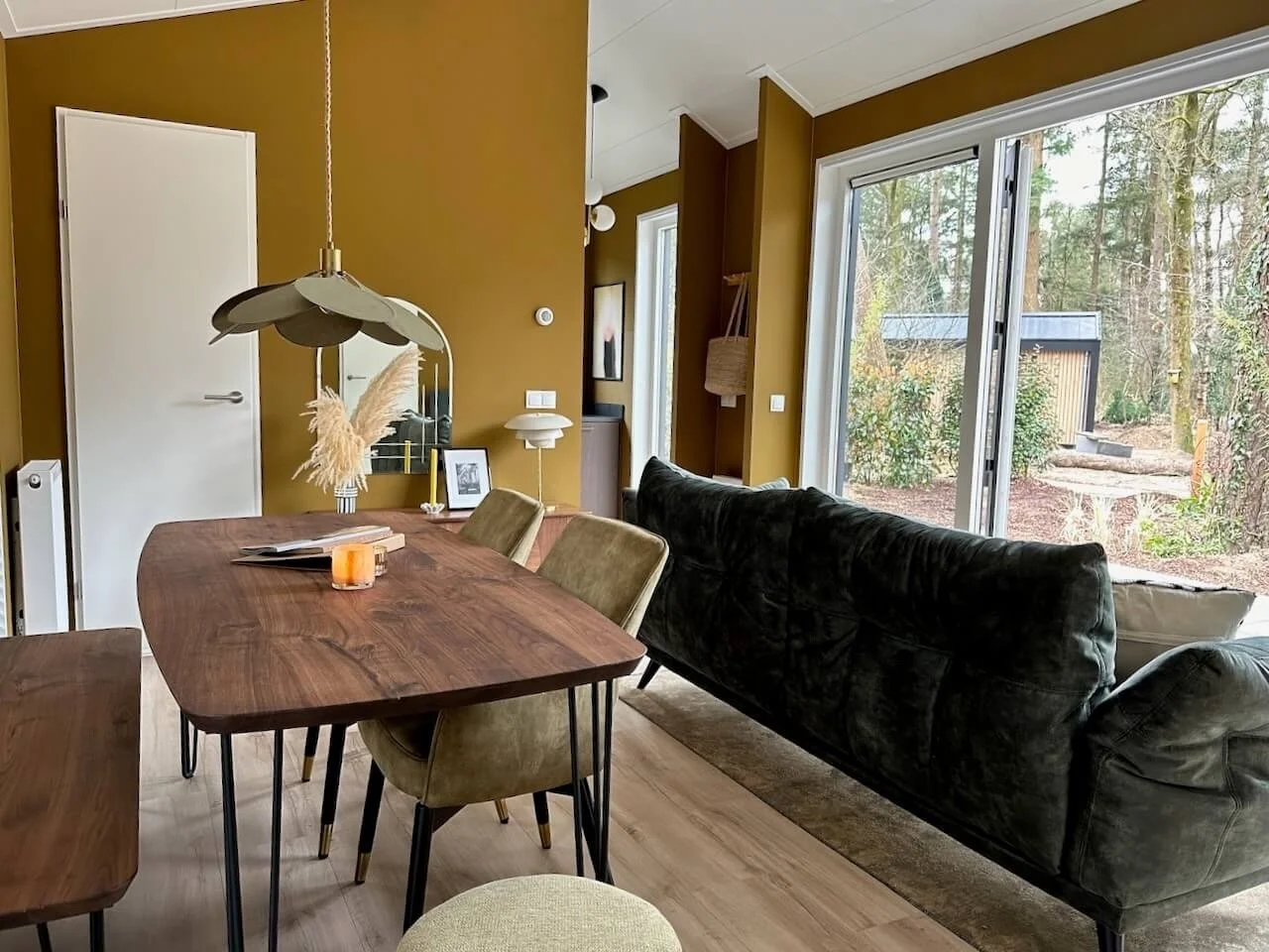 Interieur van een woonkamer met een houten eettafel, velvet stoelen, een bank, grote ramen die uitzicht bieden op een bos, en diverse decoraties.