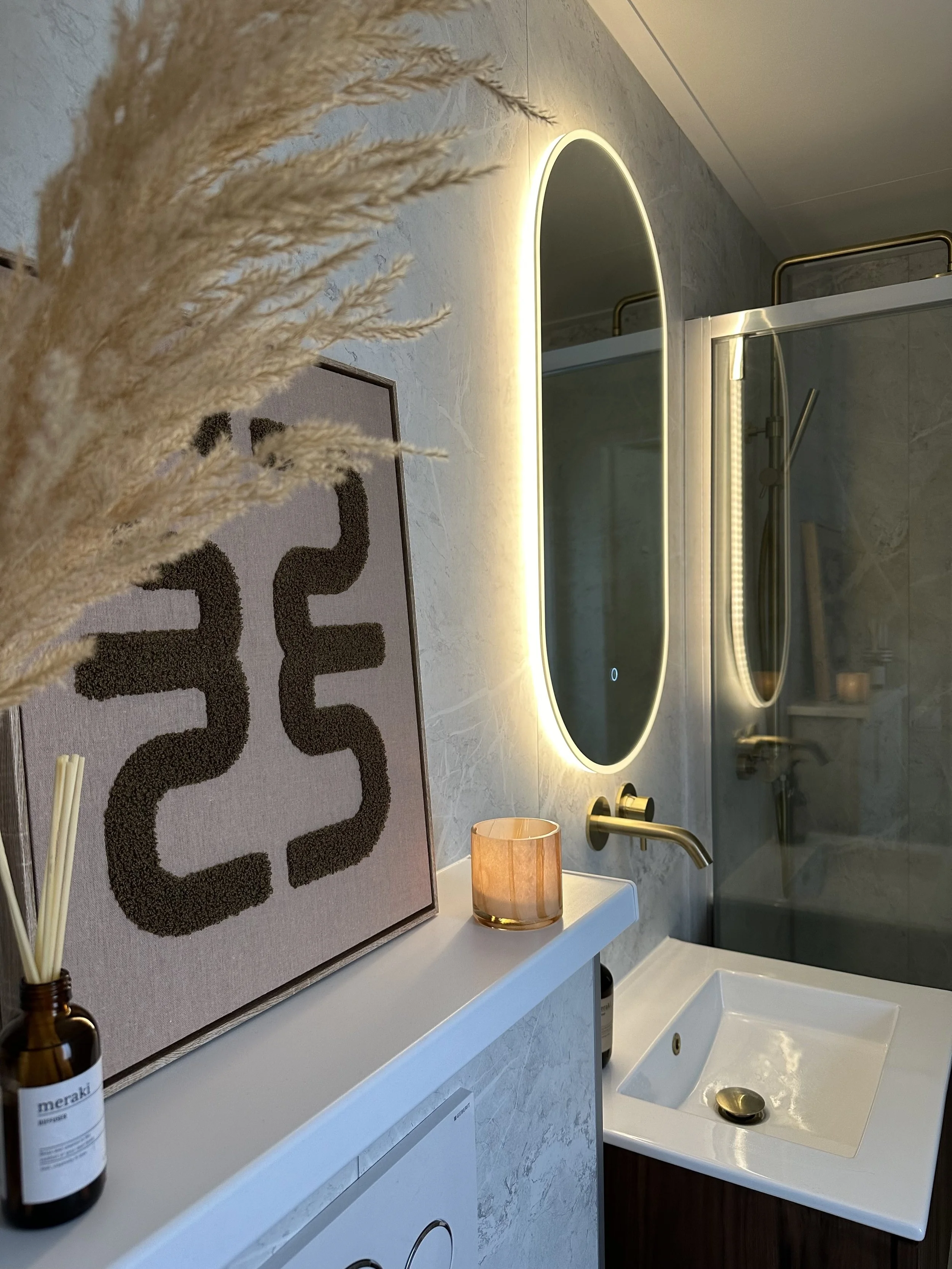 Een badkamer met een lavabo, spiegel met ingebouwd licht, een wastafel en een handdoekhouder. Decoratieve elementen zoals een doek, een lamp en een fles met lotion.