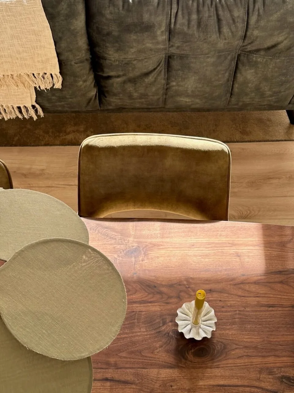 Een houten eettafel met een decoratief kaarsje in een keramische houder, omgeven door stoelen en een bank met een beige doek.