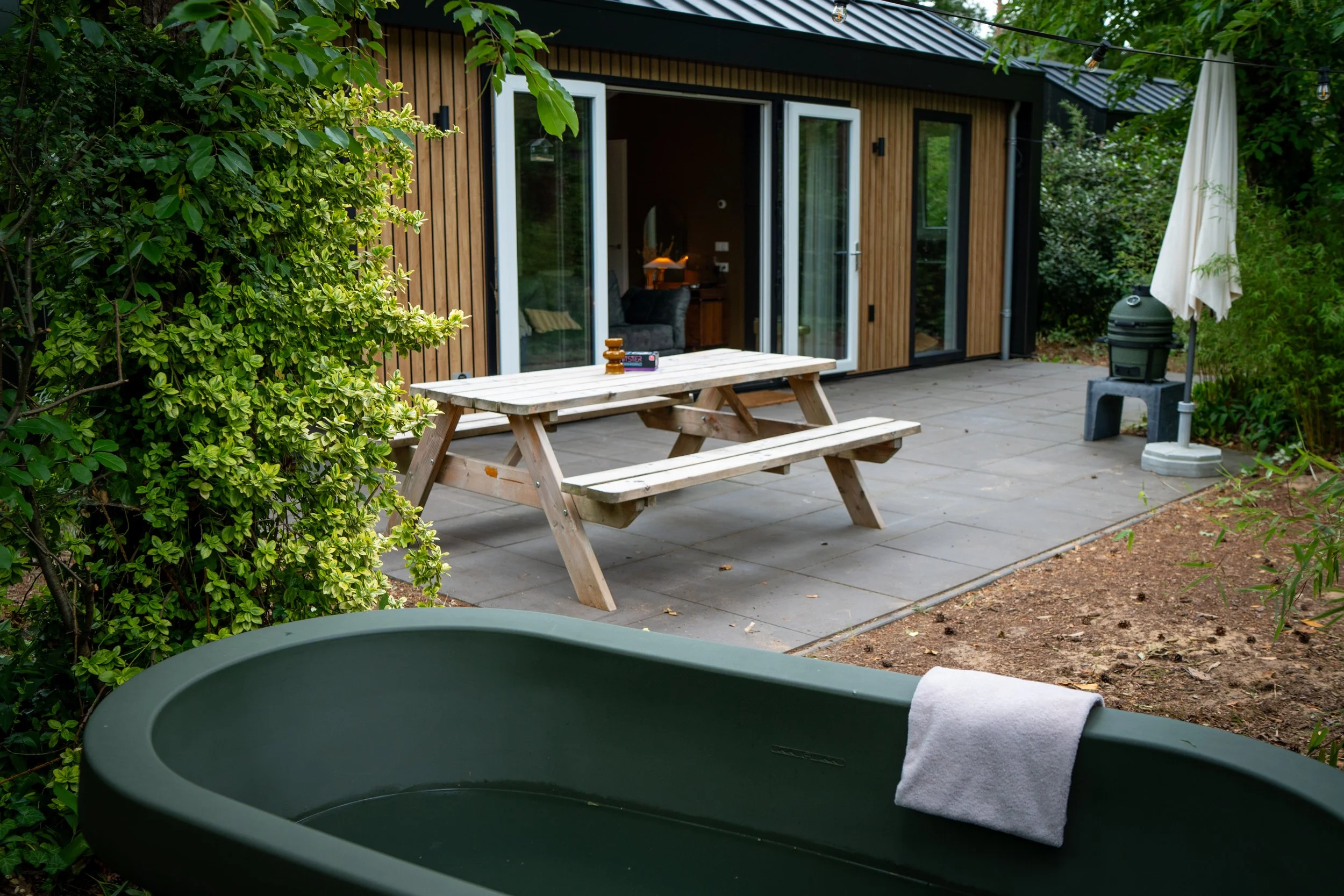 Uitzicht van een patio met een houten huis met grote glazen deuren, omgeven door groene struiken en bomen. Op de patio ligt een klassieke picknicktafel en staan lampen en een parasol.