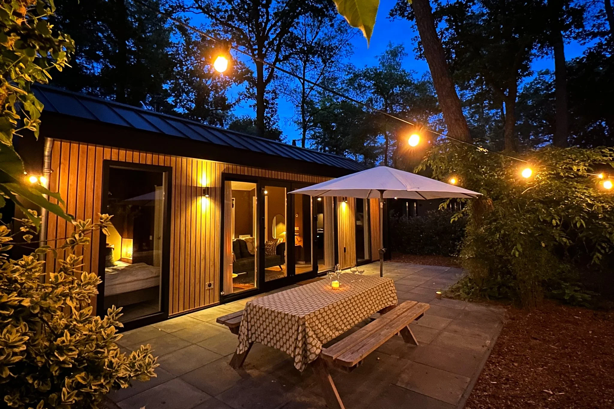 Buitenverblijf in de avond met houten huis, tafels, parasol, tuinverlichting en omringende bomen.