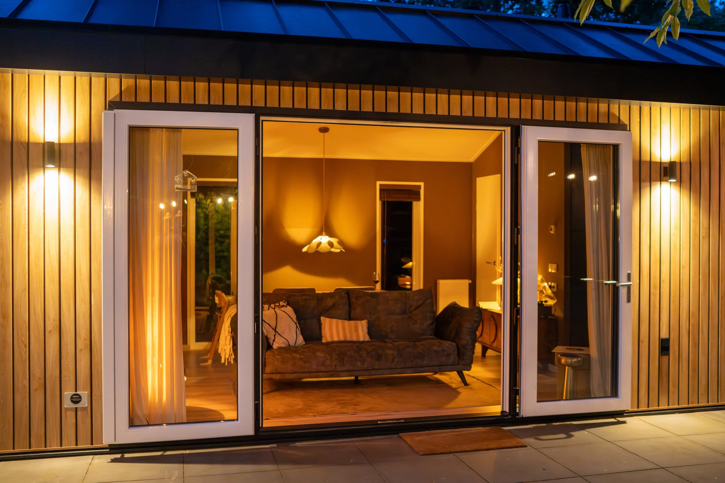 Uitzicht van een woonhuis met grote glazen schuifdeur die naar een woonkamer leidt. Binnen is een bank zichtbaar, met een lamp en decoratieve kussen, en er hangt een plafondlamp. Buiten ligt een houten vloer en een mat voor de deur.
