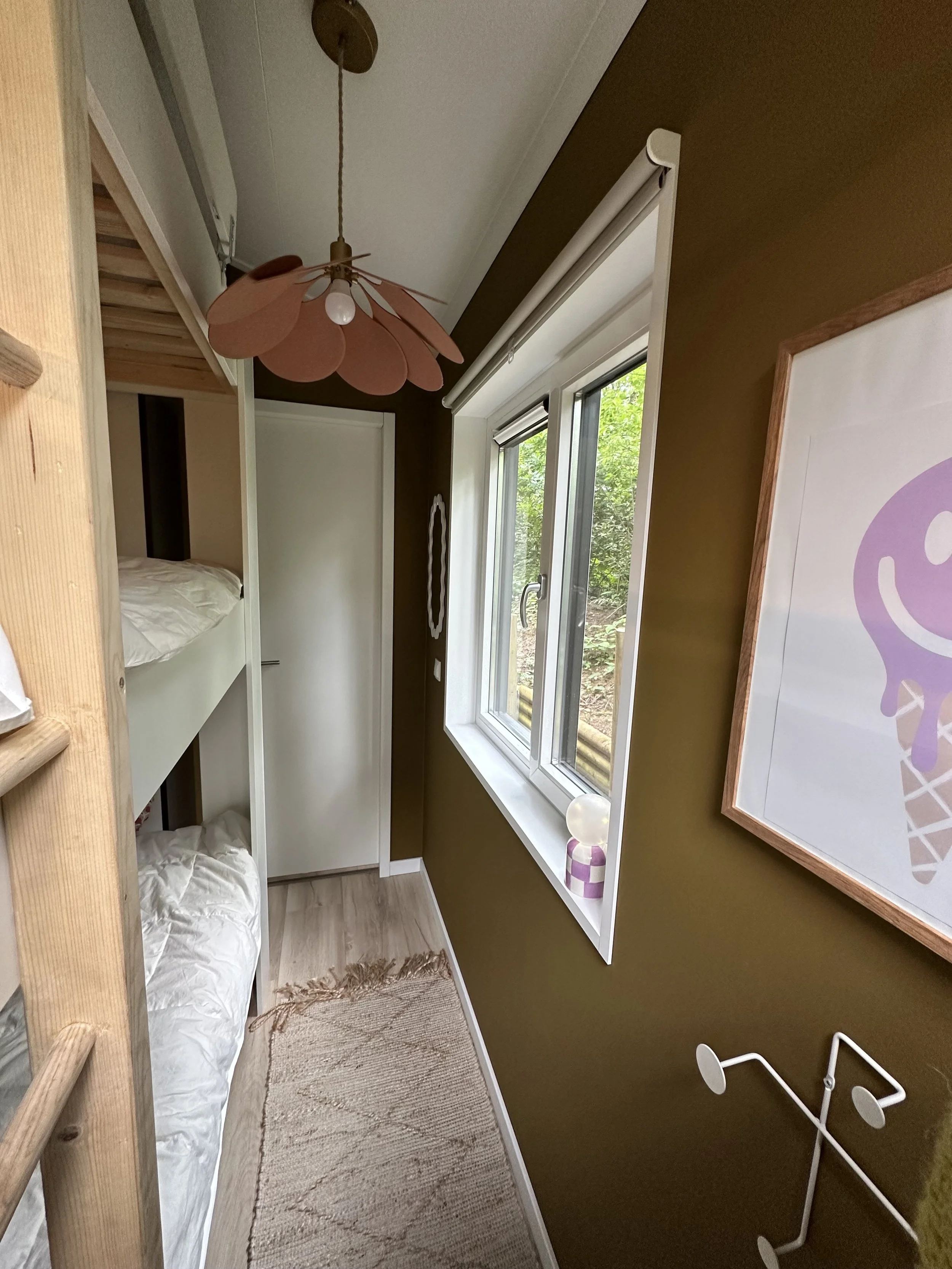 In een kleine kamer met een groenbruine muur en een houten vloer, ligt een stapelbed met witte lakens. Er staat een raam met een wit kozijn en een uitzicht op groene natuur buiten. Aan de muur hangt een poster met een paars logo en verder is er een witte plantenstandaard in de kamer.