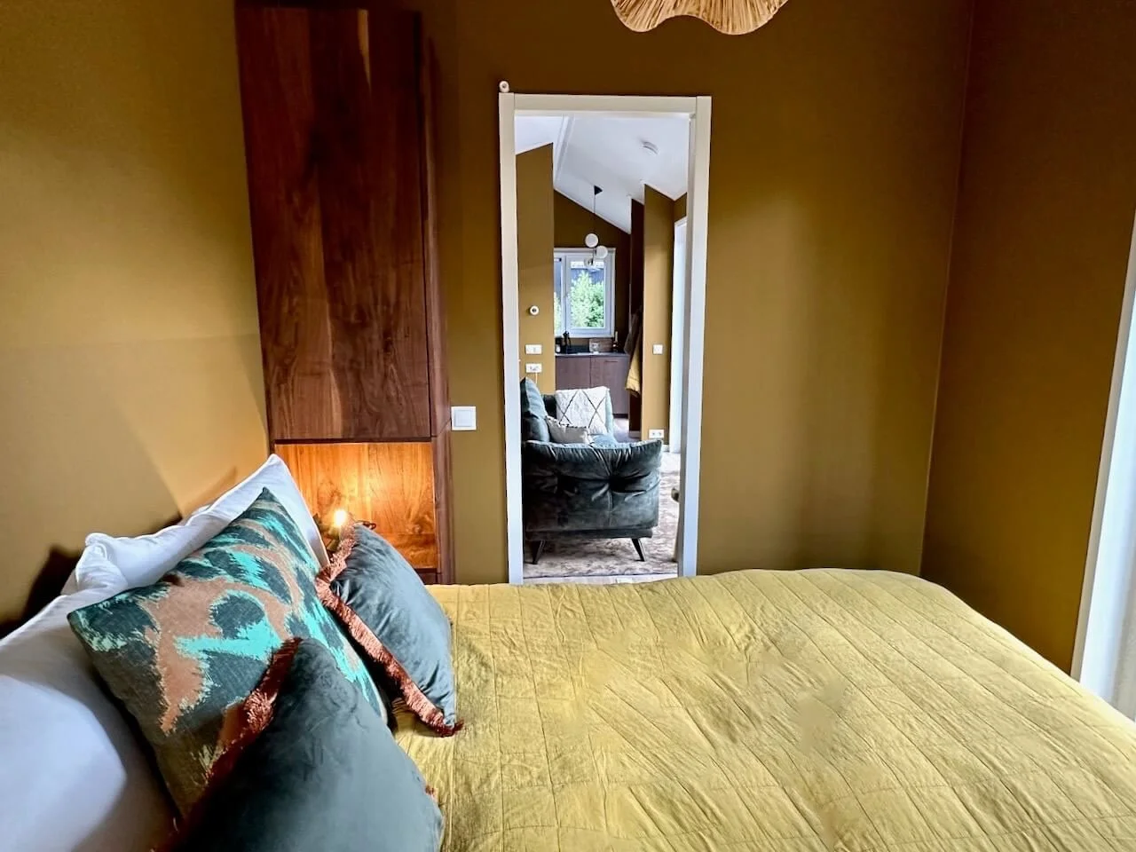 Een slaapkamer met een bed bedekt met een gele deken en decoratieve kussens, open deuropening naar een woonkamer met een zwart leren stoel en een raam in de achtergrond.