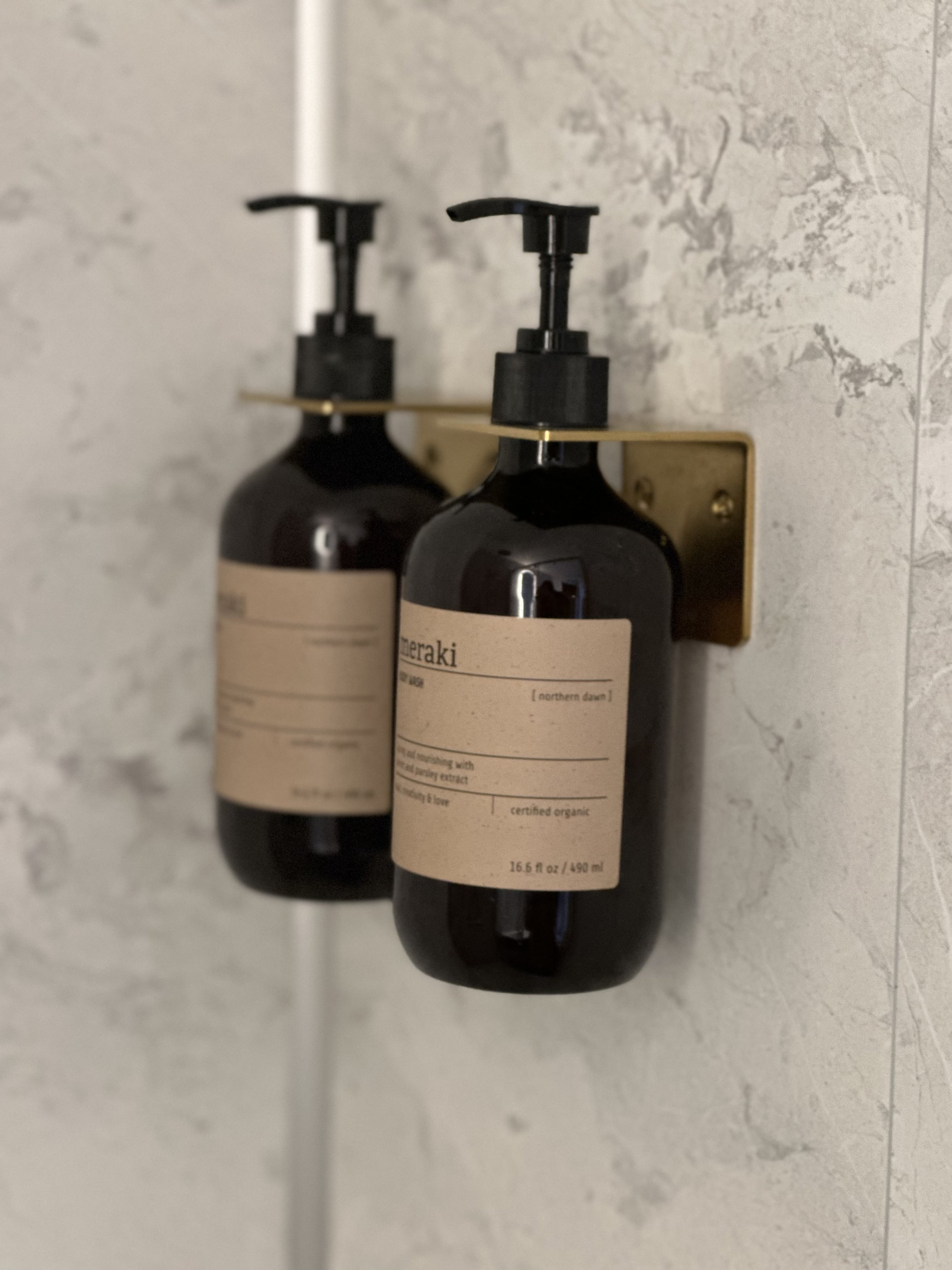 Twee zwarte flesjes met een beige label, bevestigd aan een muur met een goudkleurige houder, in een badkamer of toilet.