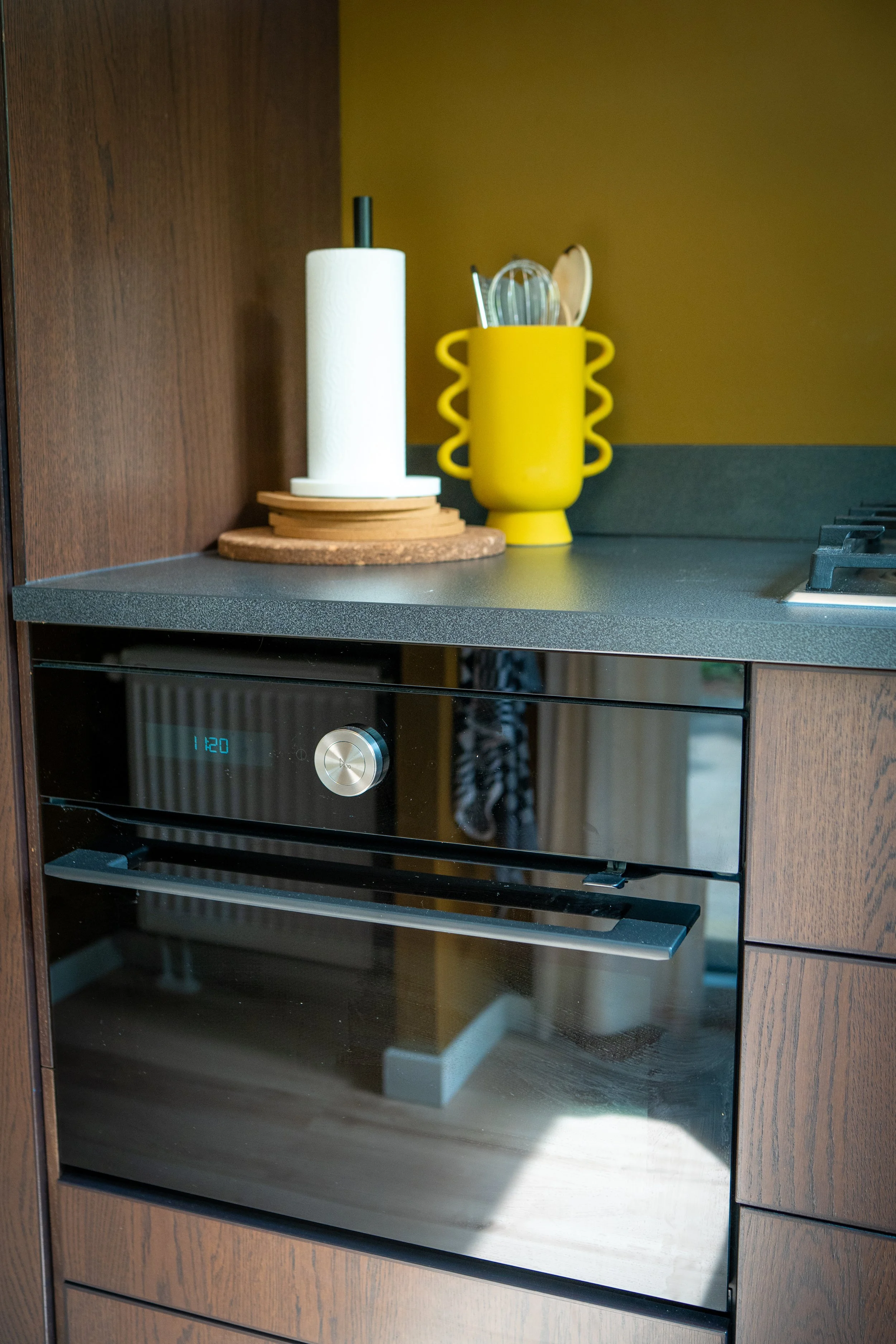 Een keuken met een oven en een zwart werkblad. Op het werkblad staan een houten snijplank, een papieren handdoekrol en een gele pot met keukengerei.