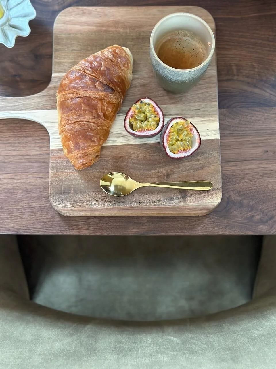 Ontbijt met croissant, passievrucht, glas thee en een gouden lepel op een houten snijplank.