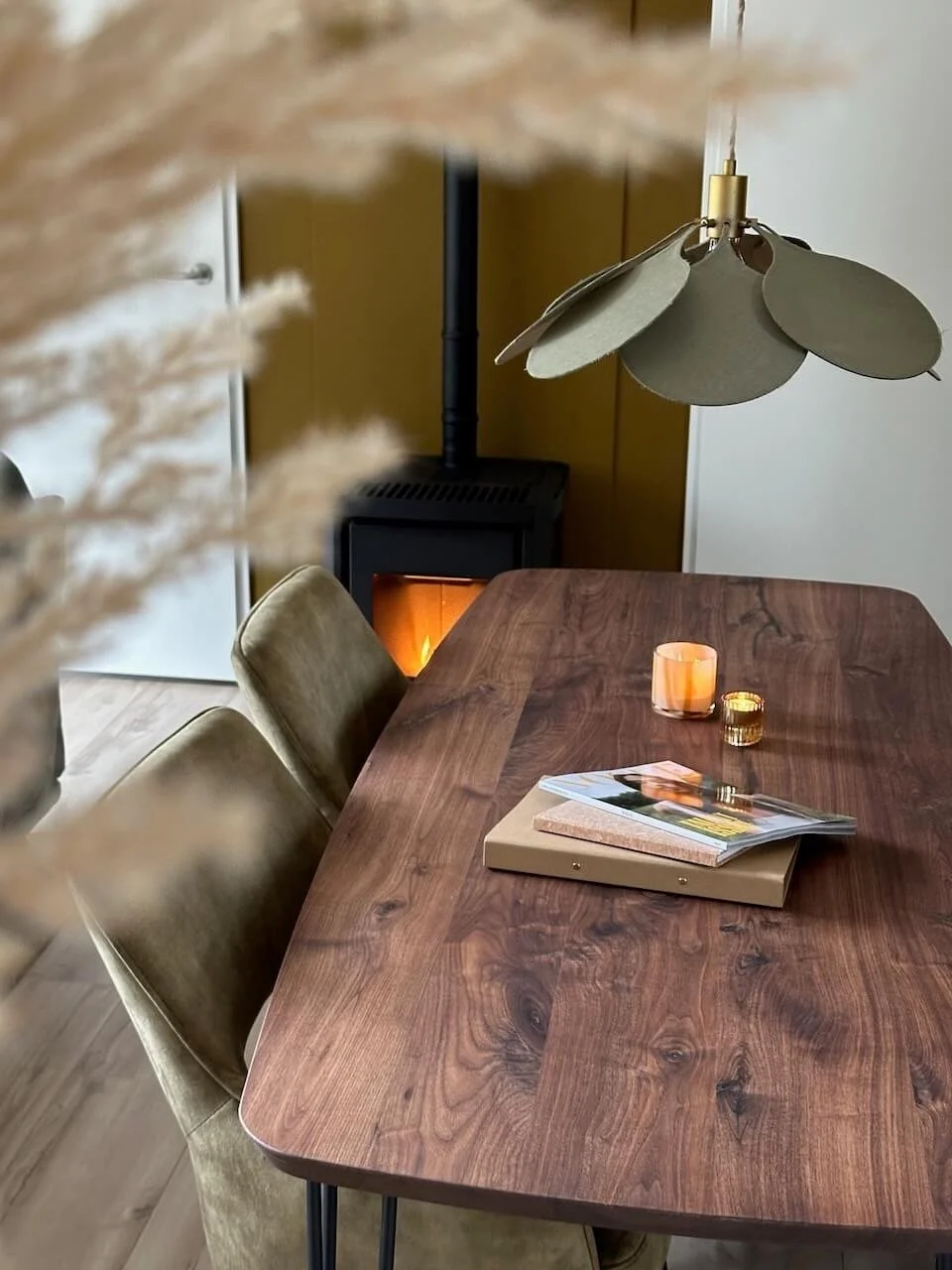 Een warme boerderij ingerichte eetkamer met een houten tafel, beige stoelen, een boek en decoratieve kaarsen, een peluw, een houtkachel en een plafondlamp.