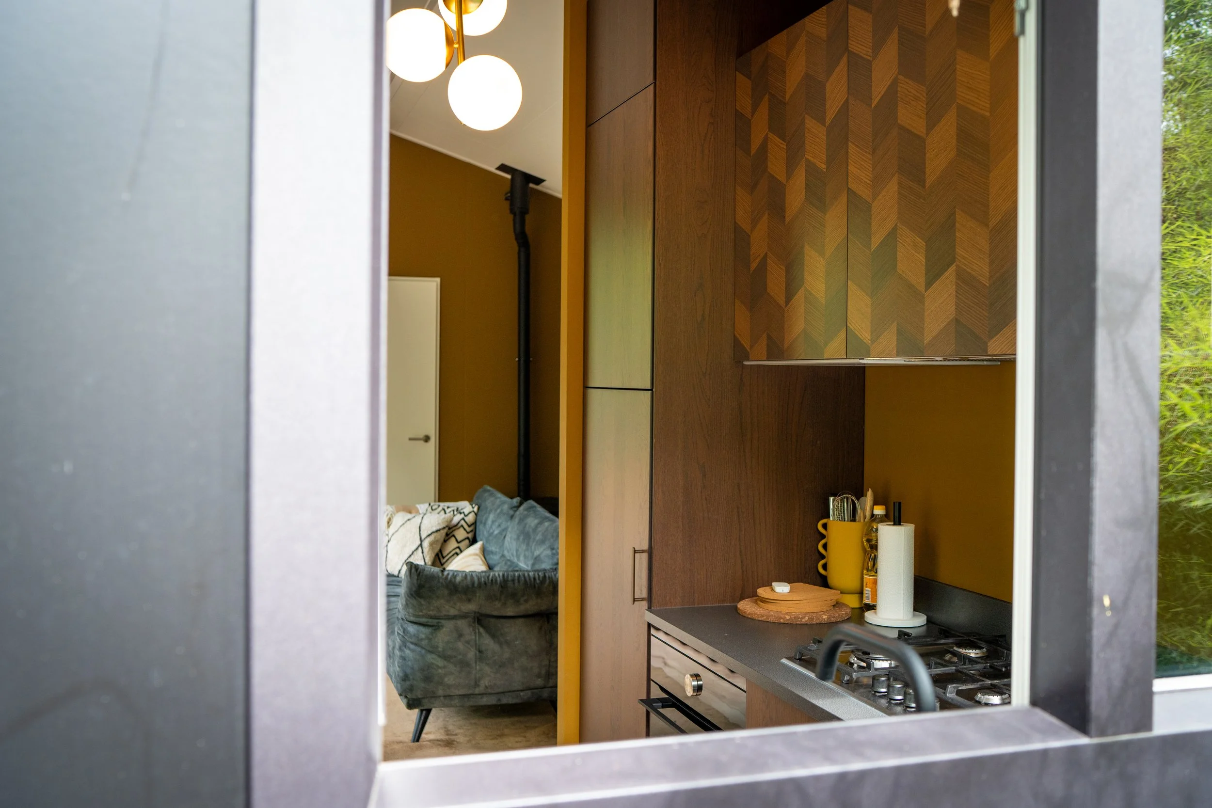 Binnenbeeld van een moderne keuken met een gaskookplaat, een keukenblok met accessoires, een woonkamer met een bank en een muur met houten panelen, gezien via een raam of opening.