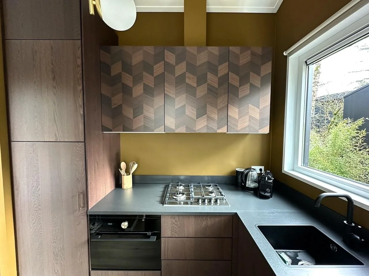 Kleine keuken met houten kasten, gasfornuis, gootsteen, en apparaten zoals waterkoker en koffiezetapparaat bij raam met uitzicht op tuin.